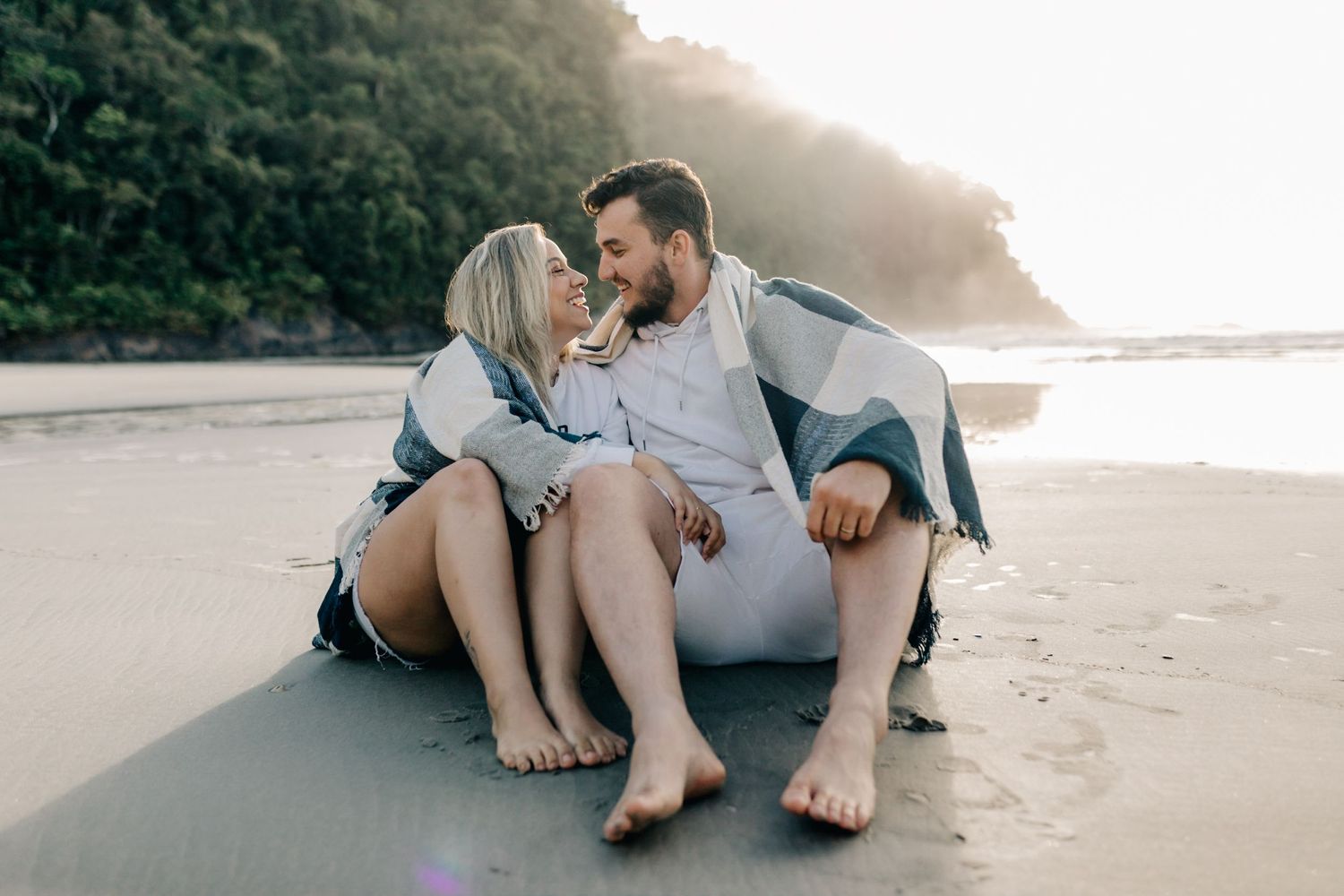 Couple cuddling on sandy beach with cliffside and ocean in background during golden hour.