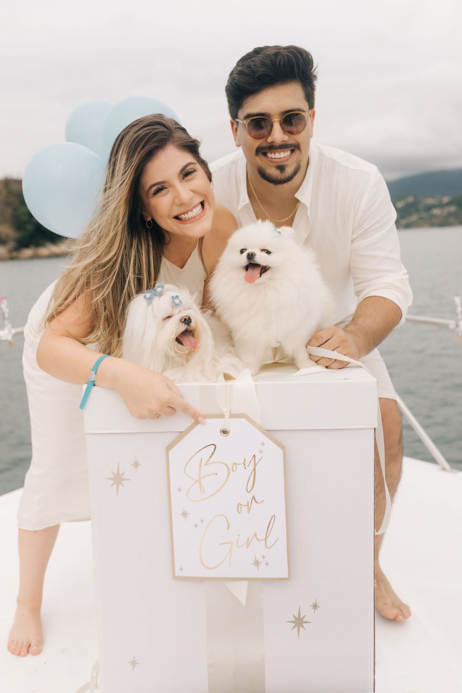 A couple and their two white dogs pose together on a boat for a gender reveal announcement.