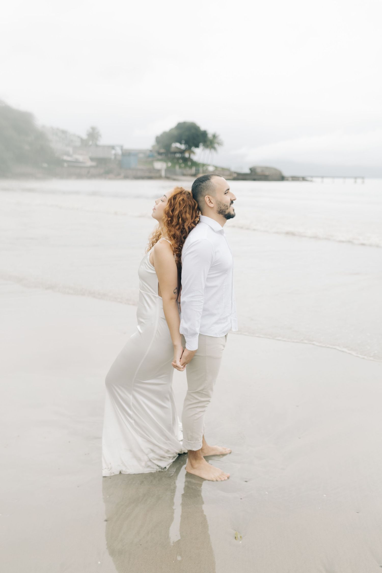 Couple in white standing barefoot on wet sand at a misty beach.