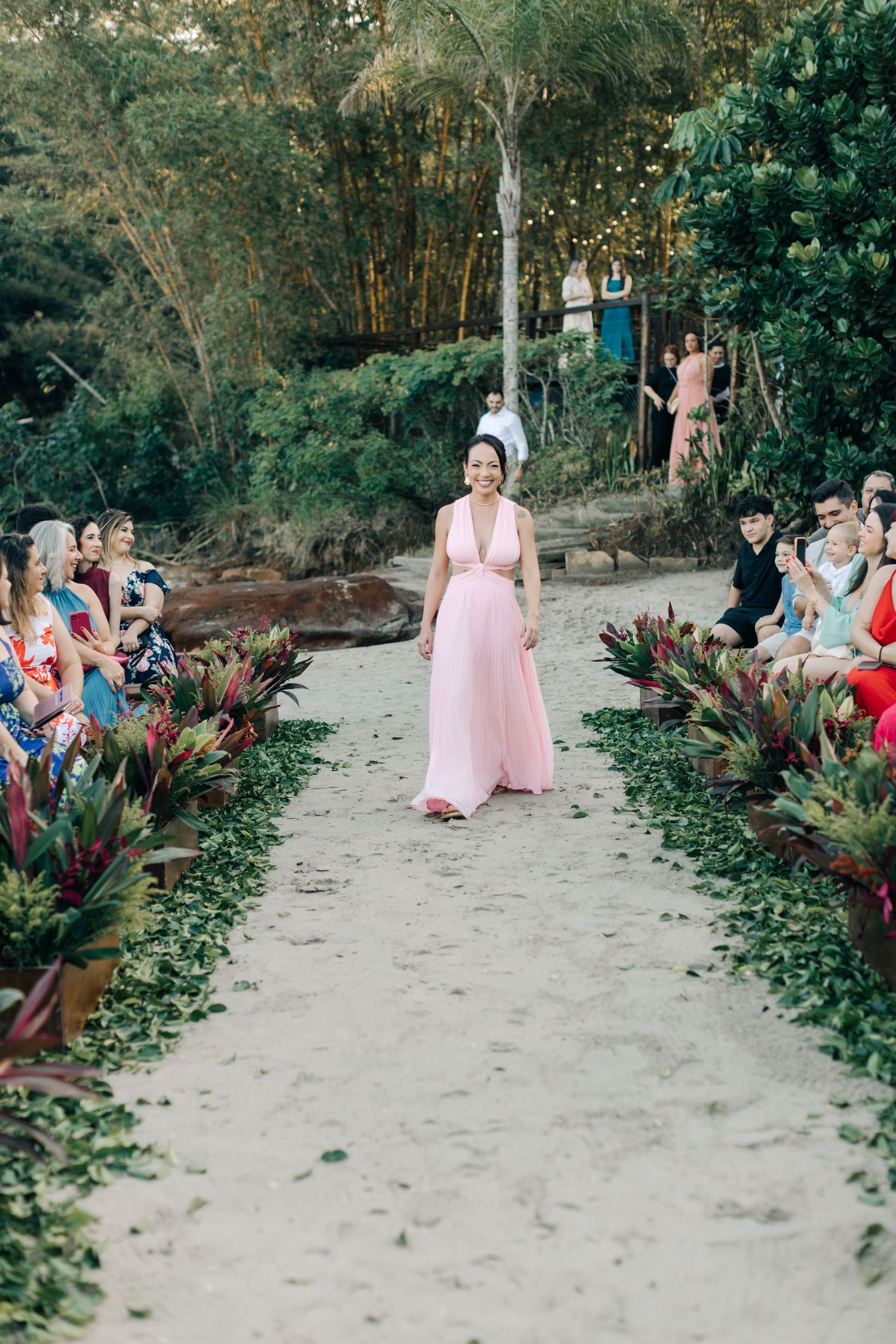 Bridesmaid in soft pink gown walks down tropical garden path at outdoor wedding.