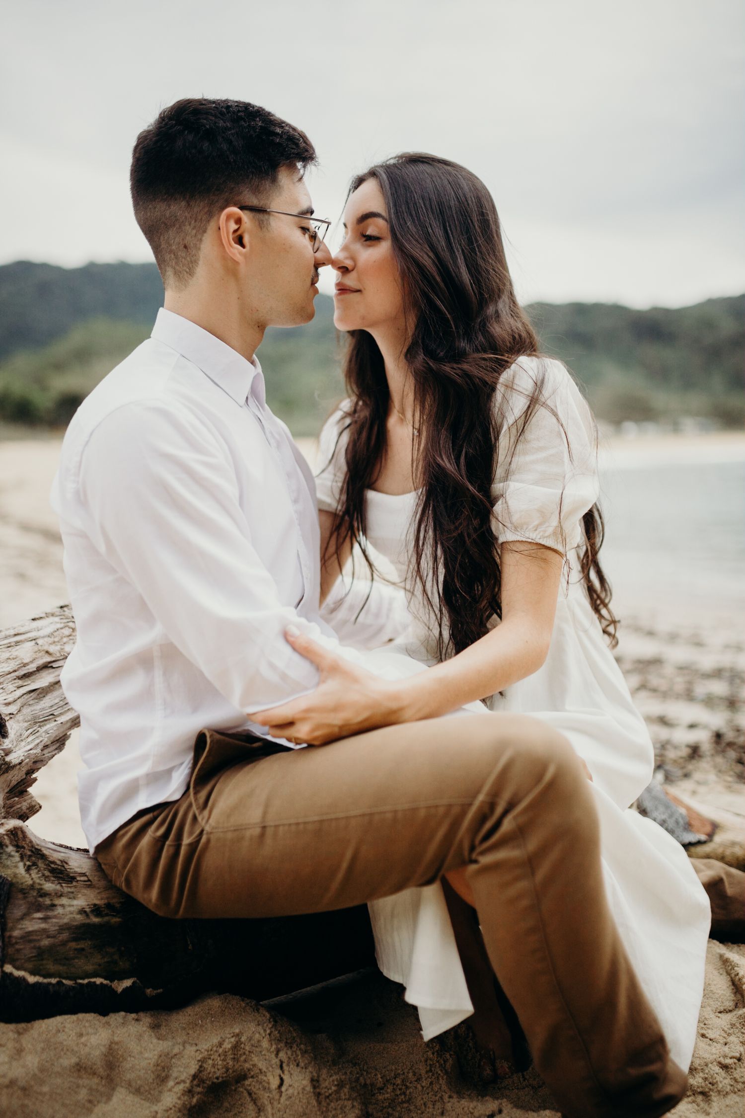 Couple shares tender moments while sitting on driftwood at the beach.