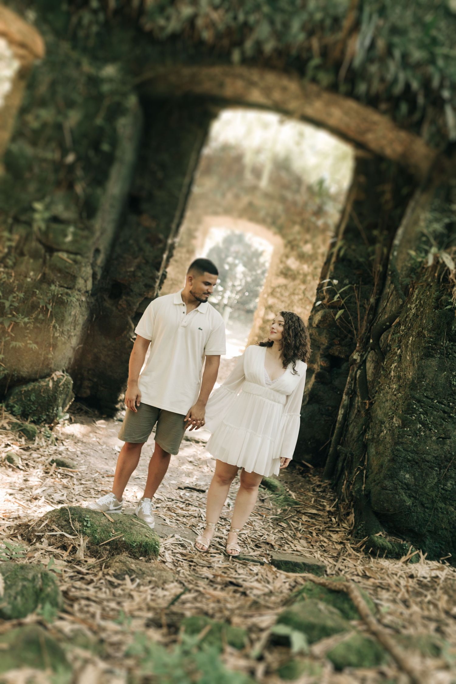 Artistic forest photoshoot featuring couple walking through stone ruins in matching white attire.