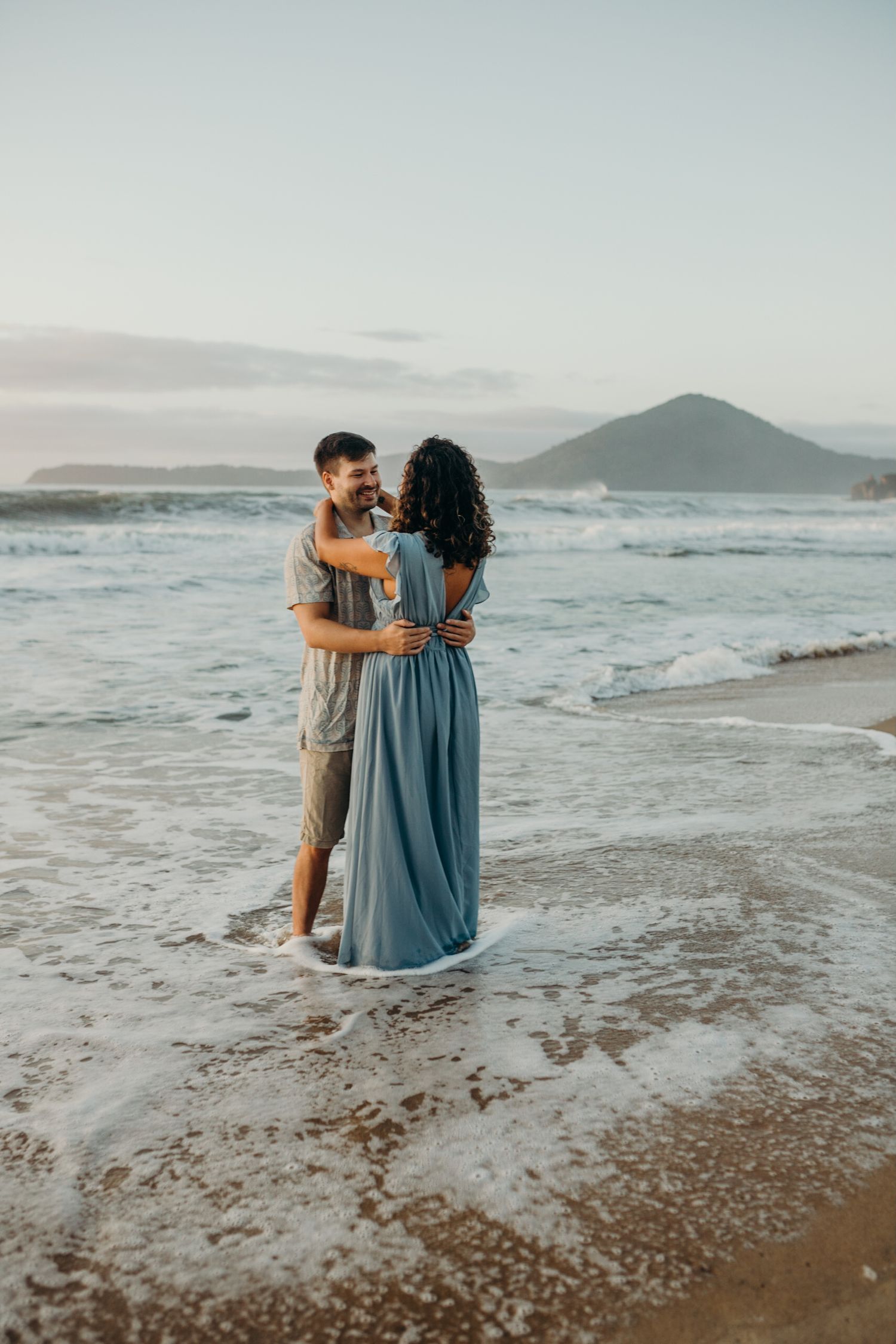 Romantic couple sitting in shallow ocean waves during beach photoshoot at sunset.