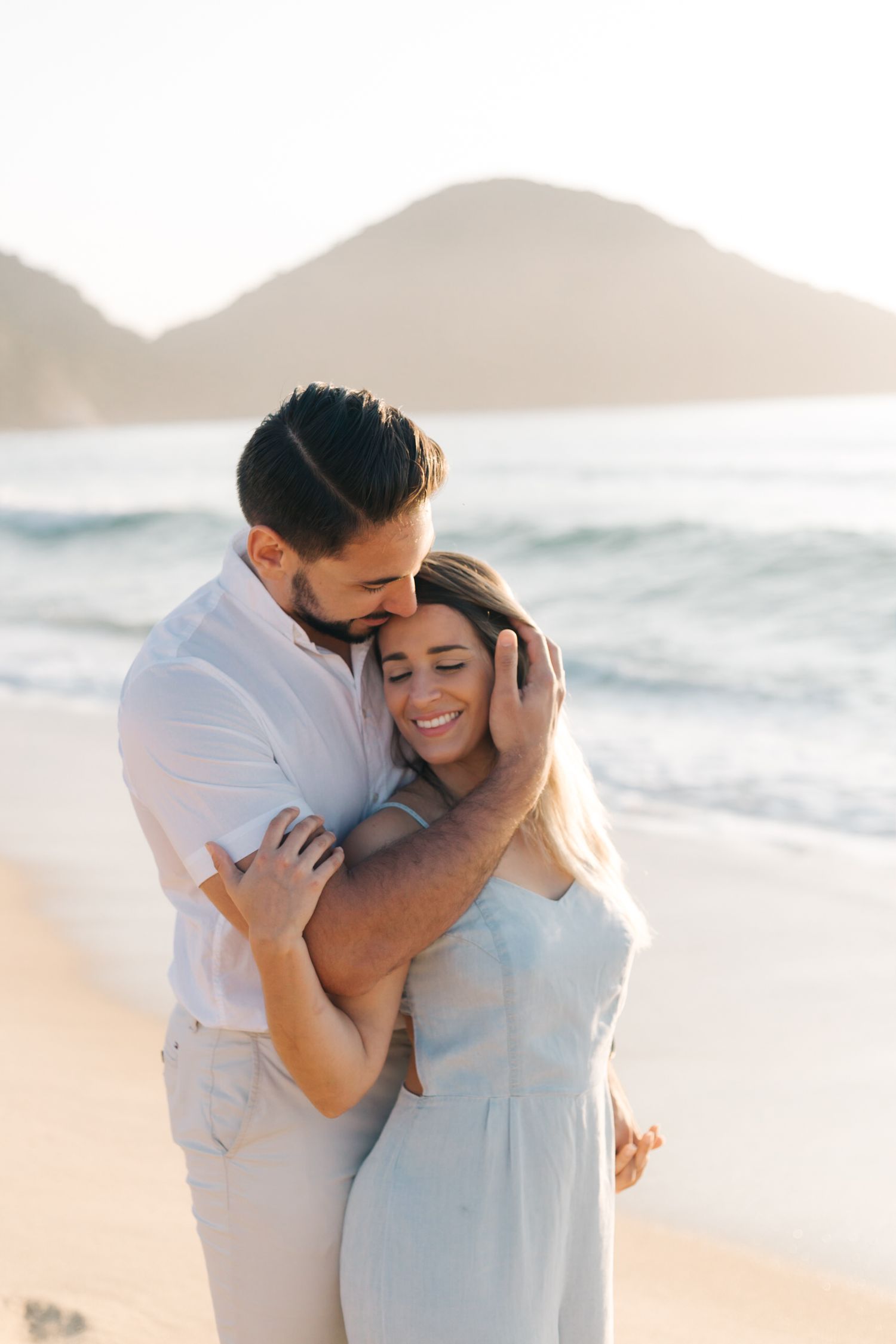 Tender embrace between couple in white outfits on scenic beach during golden hour.