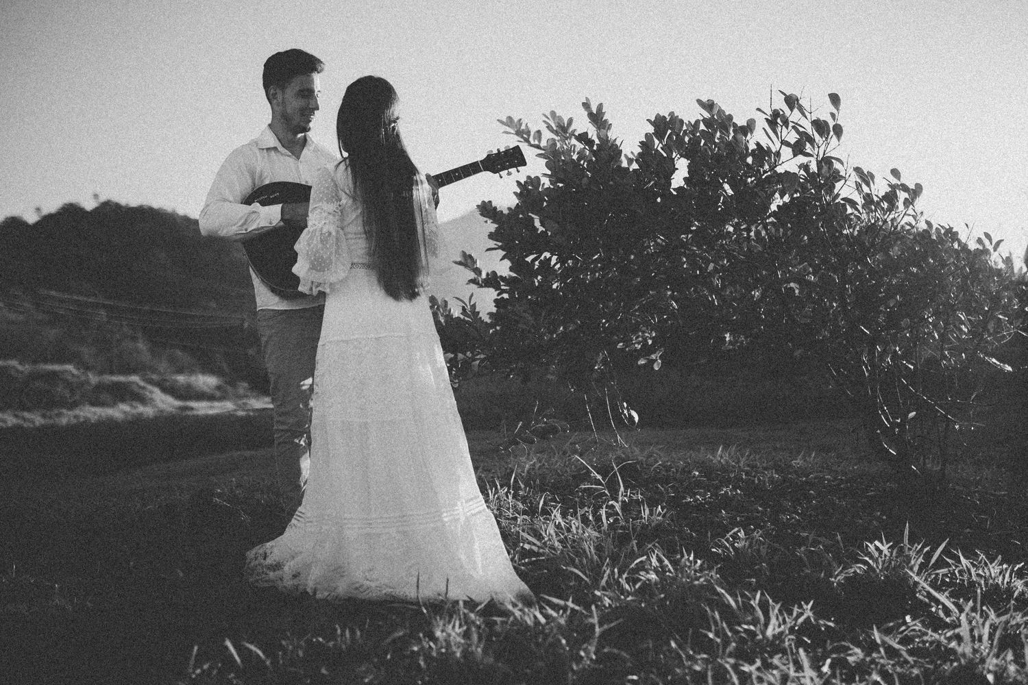 Black and white photo of newlyweds standing outdoors with guitar during sunset.