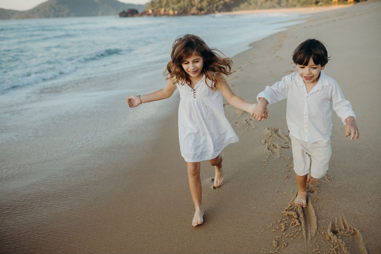 Two figures in white outfits playing and running on a scenic beach with mountains and ocean views.