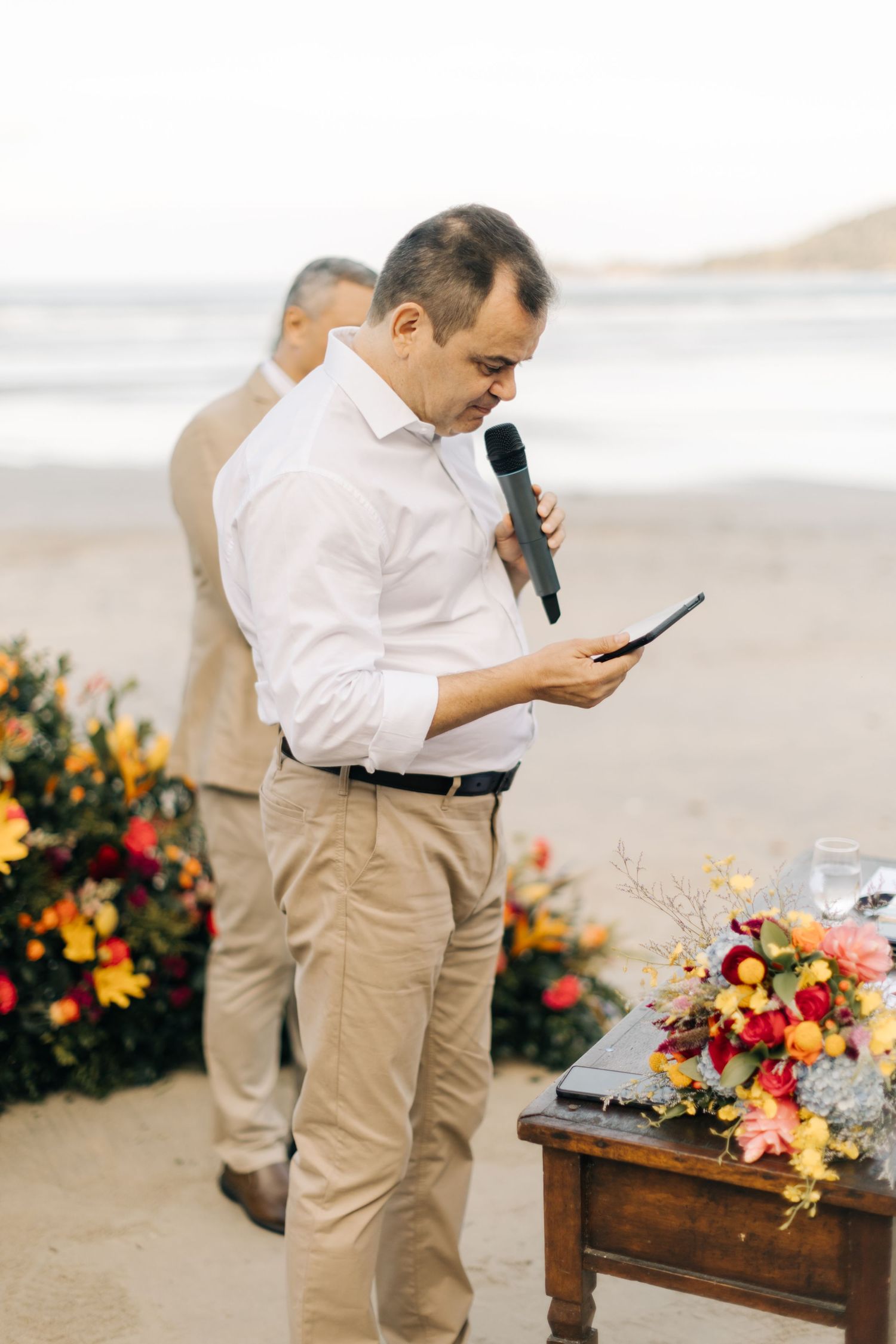 Wedding officiant in white shirt and khakis speaking at a beach ceremony with floral decorations.