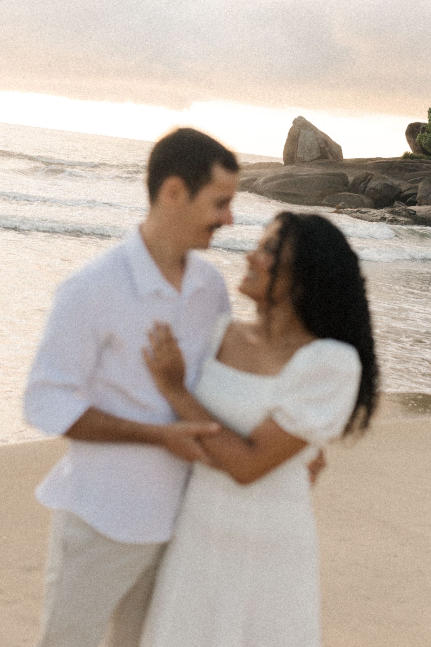 A couple shares a romantic moment on a sandy beach during sunset with ocean waves and cliffs in the background.