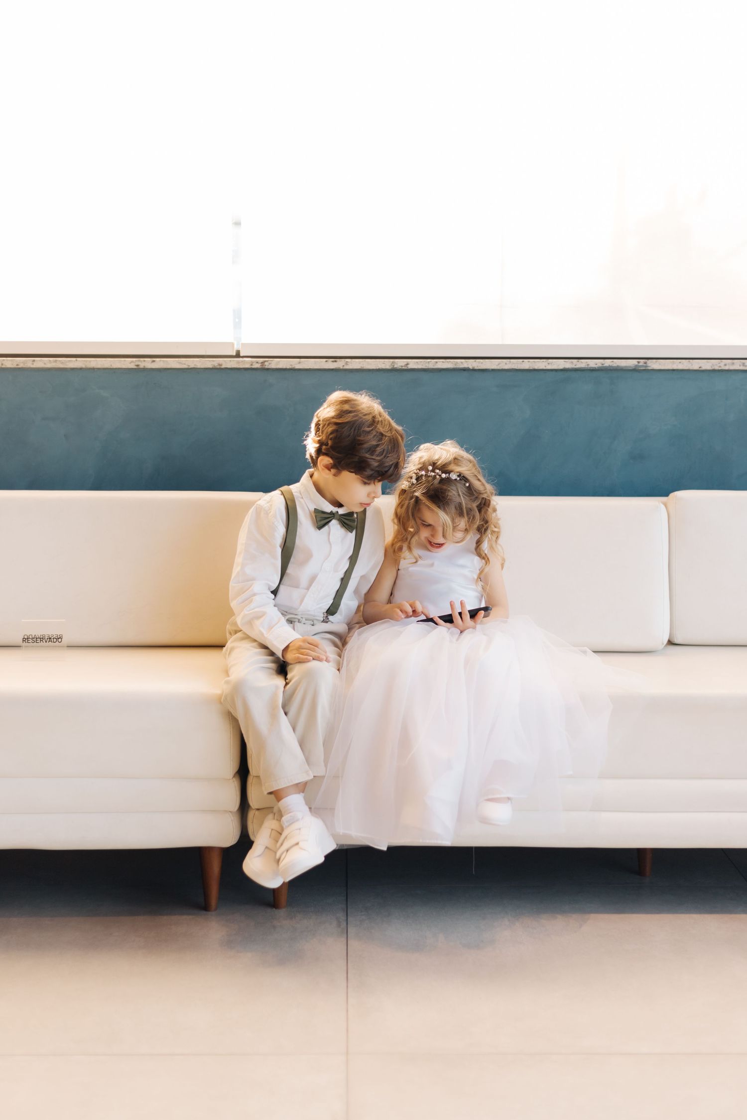 Two children in formal white attire sitting together on a modern couch.