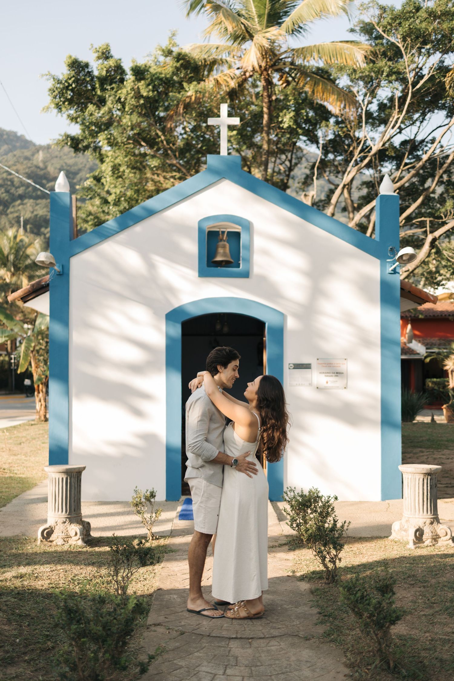 A couple shares a romantic moment in front of a charming white chapel with blue trim and palm trees in the tropical setting.