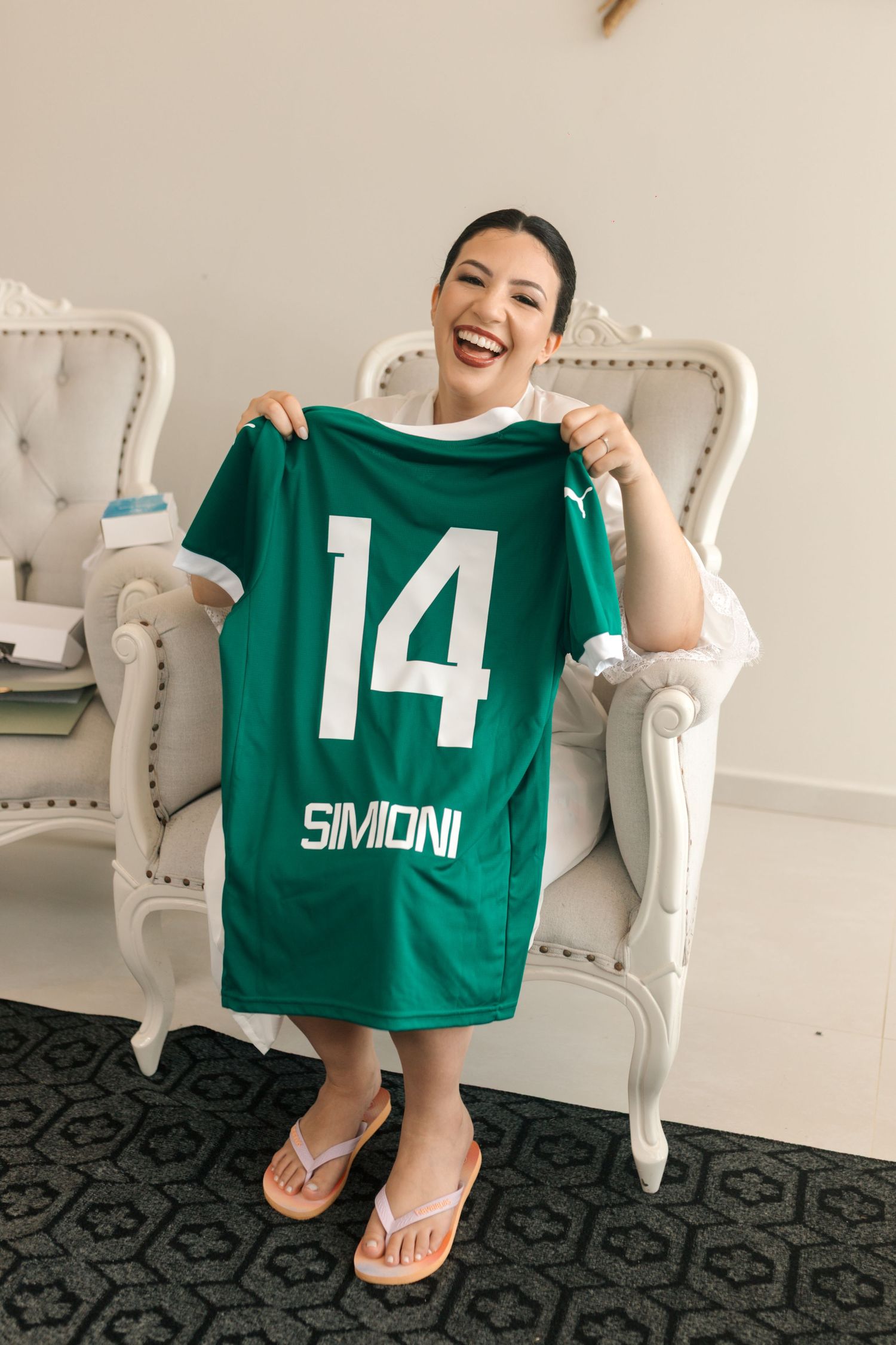 Person wearing white robe displays green sports jersey number 14 while sitting on vintage chair.