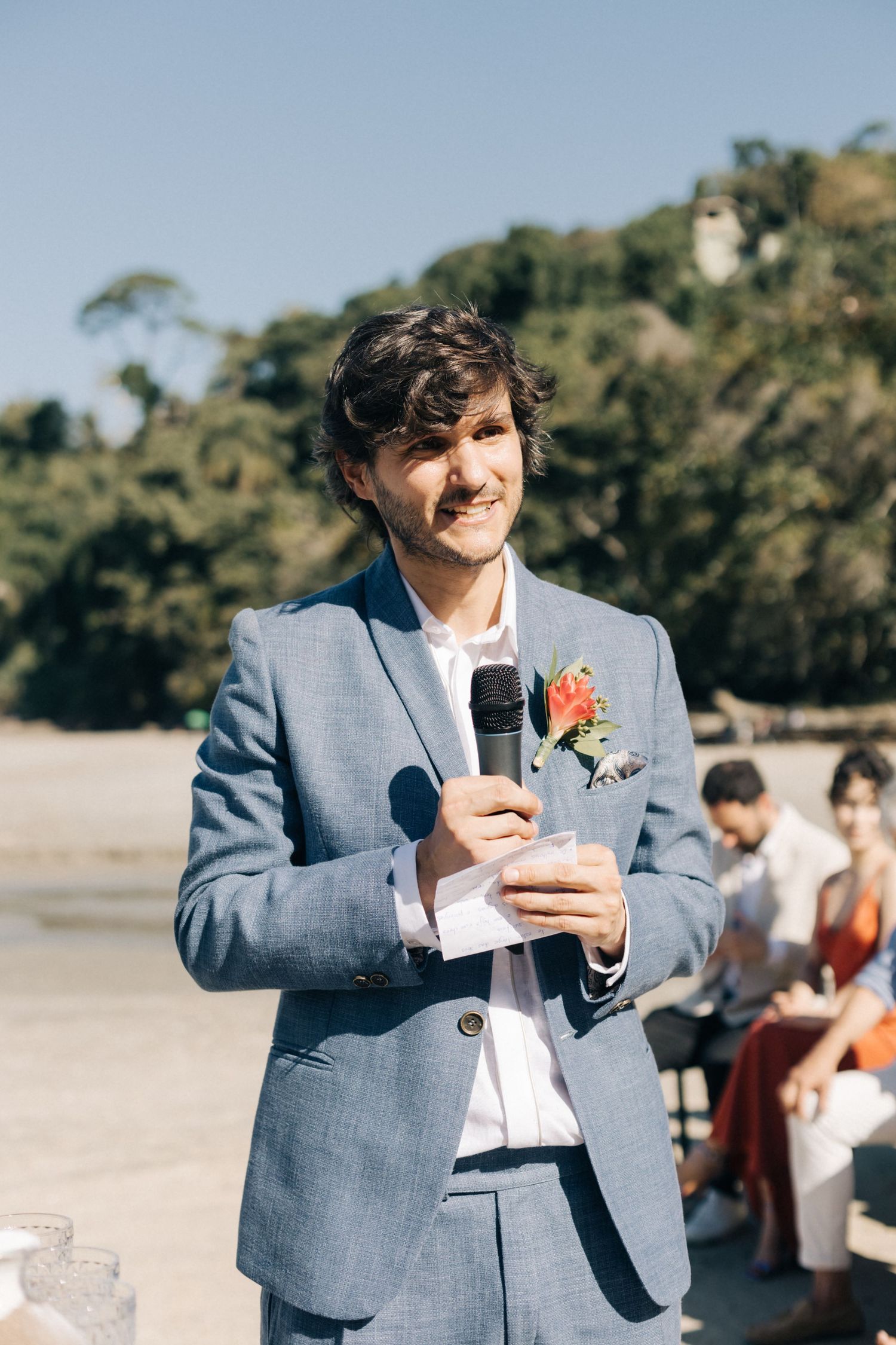 A person in a light blue suit delivers a speech during an outdoor beach wedding.