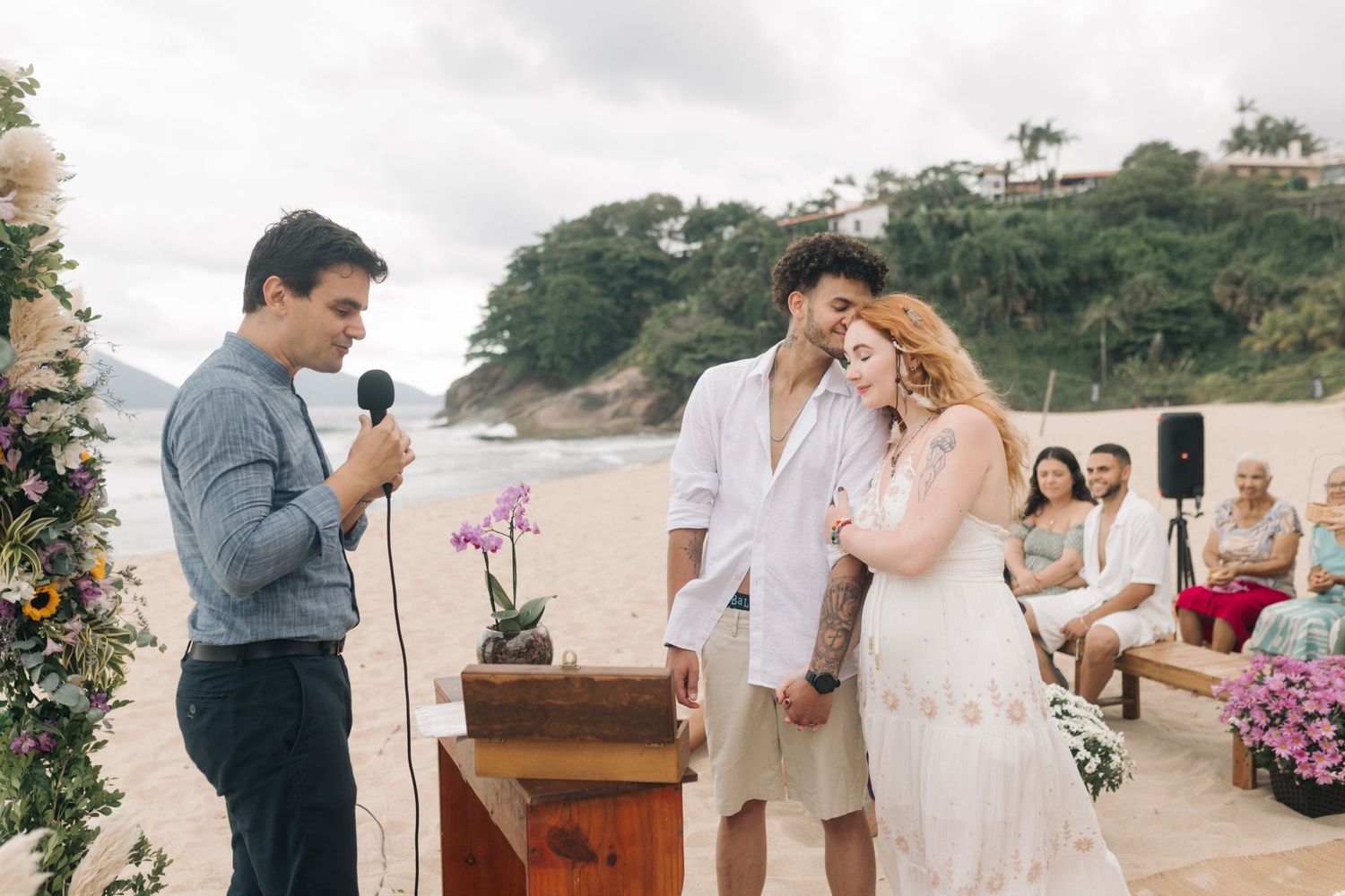 A beachside wedding ceremony with an officiant speaking to the couple under cloudy skies.