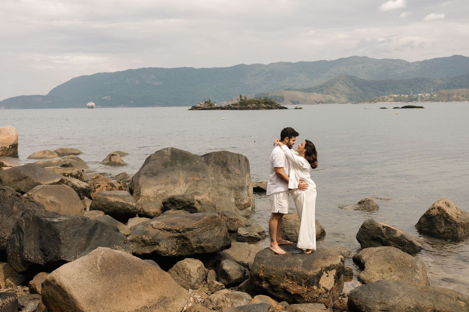 A couple shares a romantic moment on rocky coastal shoreline with mountains and ocean in the background.