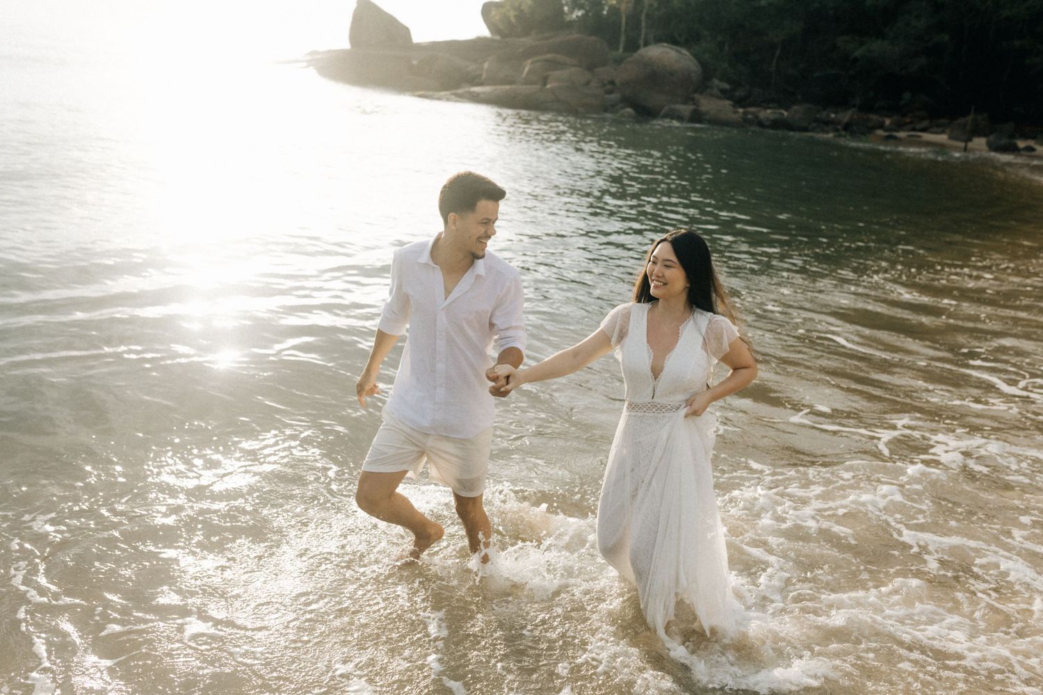 Playful couple splashing in shallow water wearing white beach attire.