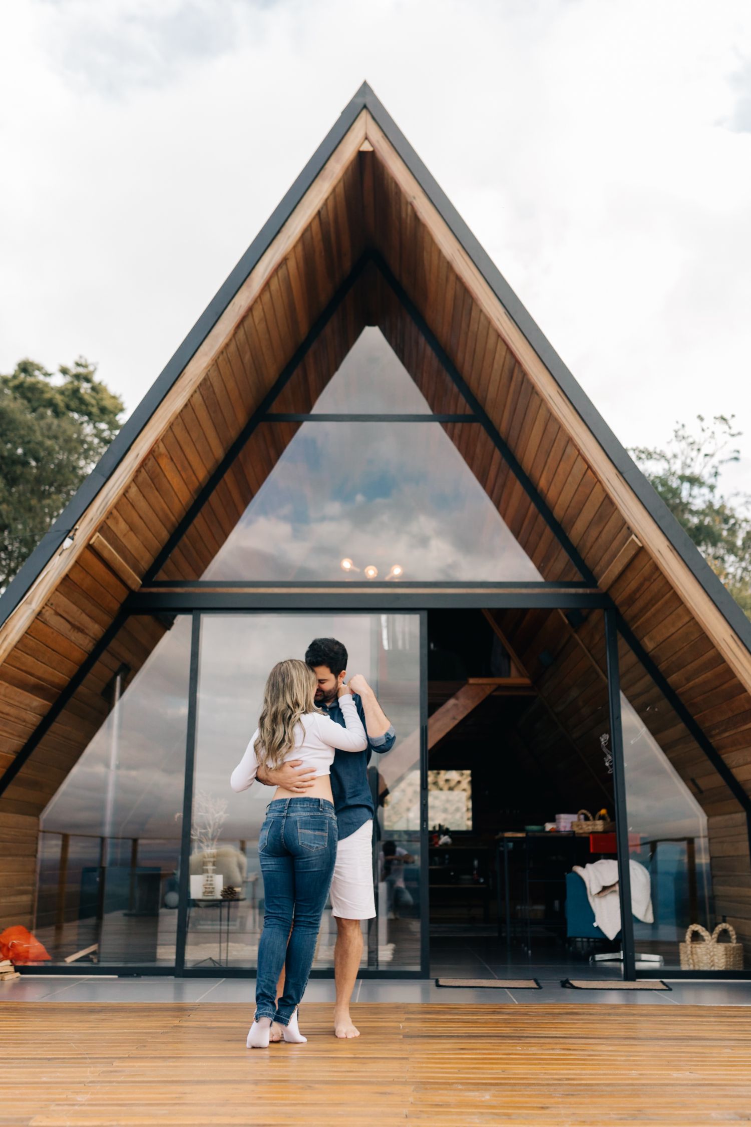 A modern A-frame cabin with glass walls and wooden accents on a deck against cloudy skies.