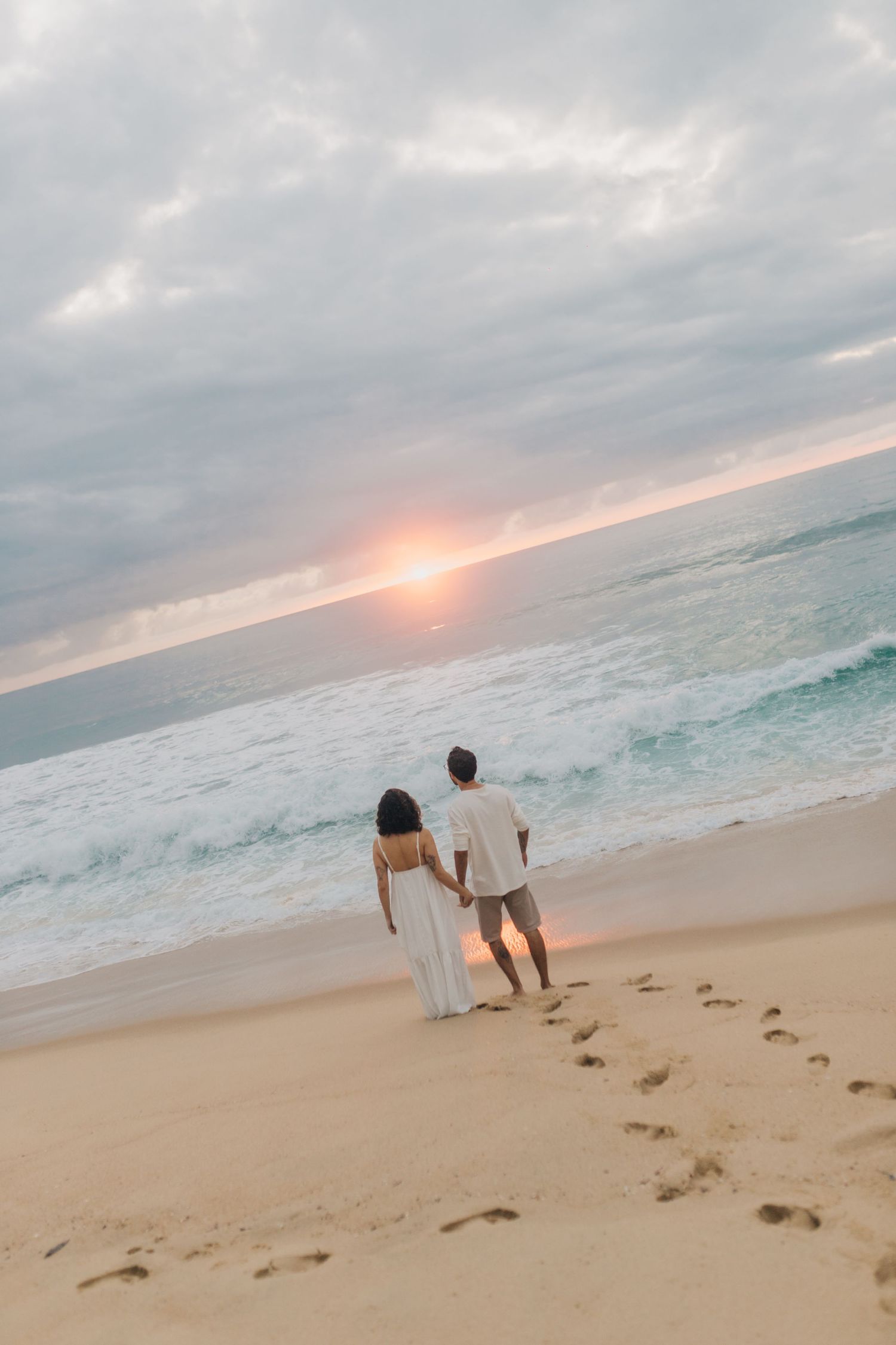 A couple holds hands while walking on a sandy beach at sunset leaving footprints in their wake.