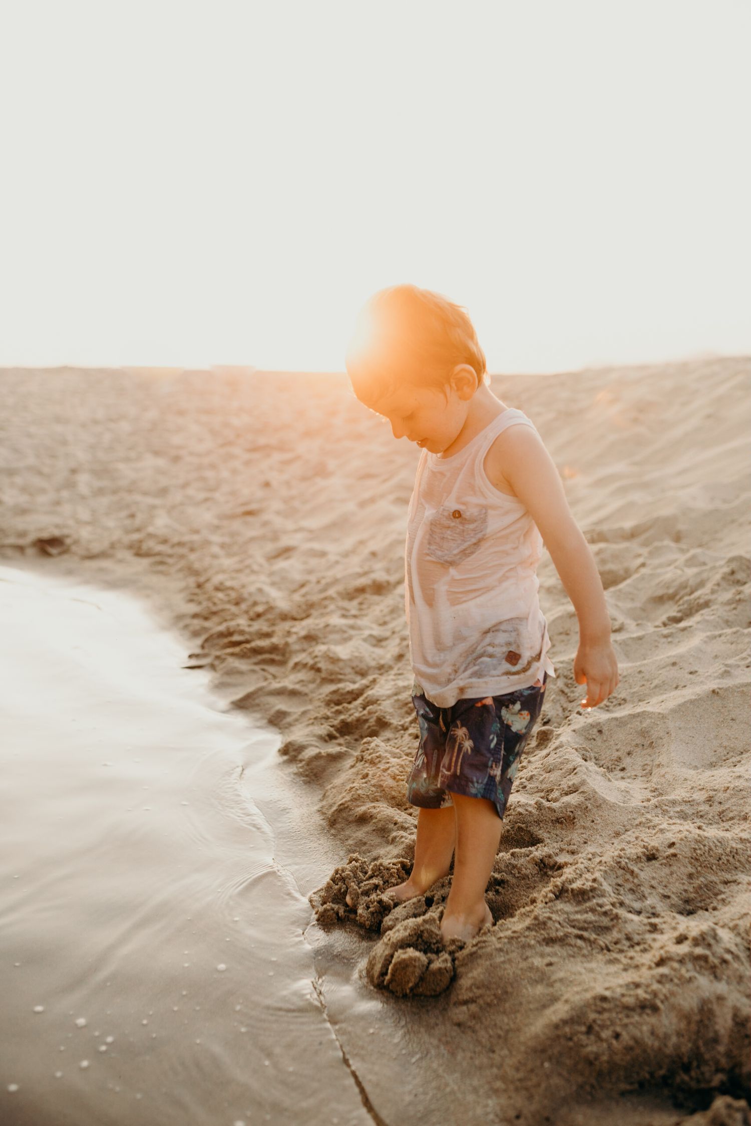 Child standing at water's edge on beach during golden hour sunset.