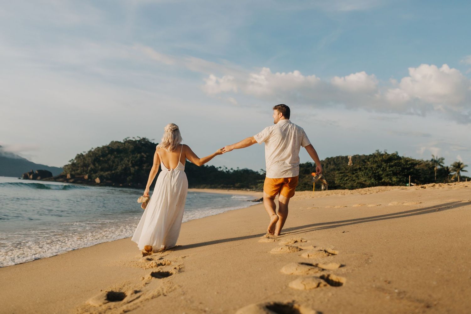 Playful couple running along shoreline leaving footprints in wet sand during beach photoshoot.