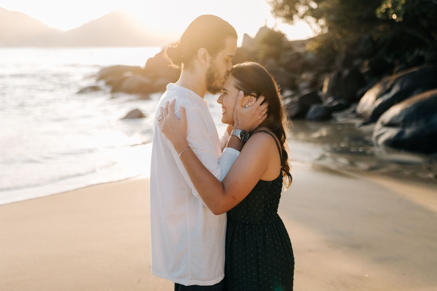 Couple sharing romantic moment on sandy beach with dramatic coastal scenery.