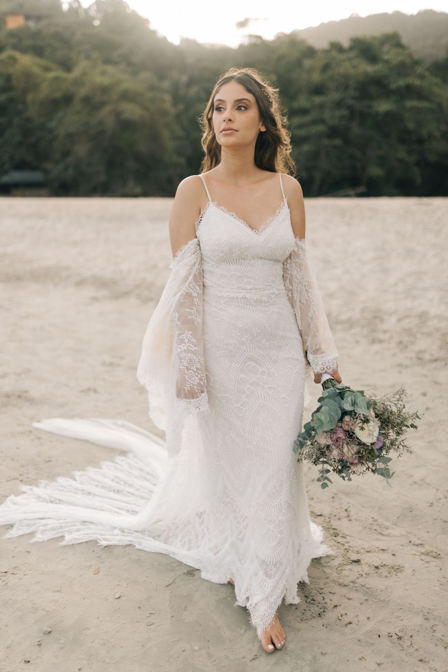 Bride in elegant lace wedding dress holds bouquet while standing barefoot on beach at sunset.