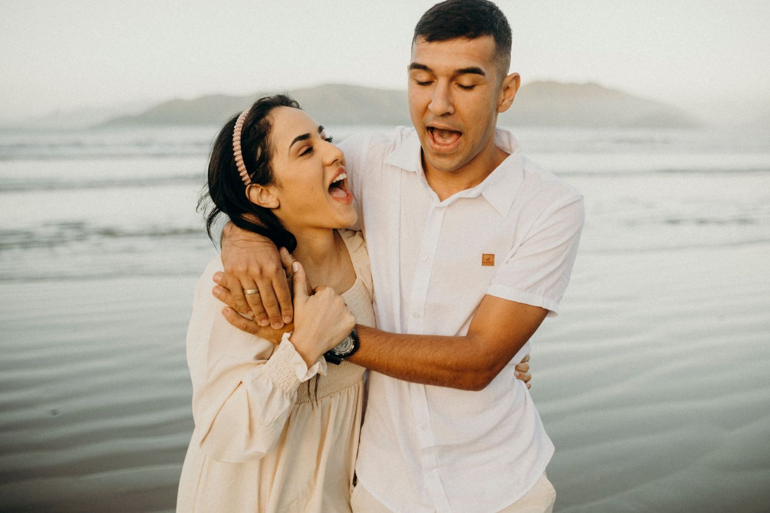 Playful couple embracing and making funny faces on the beach with mountains in background.