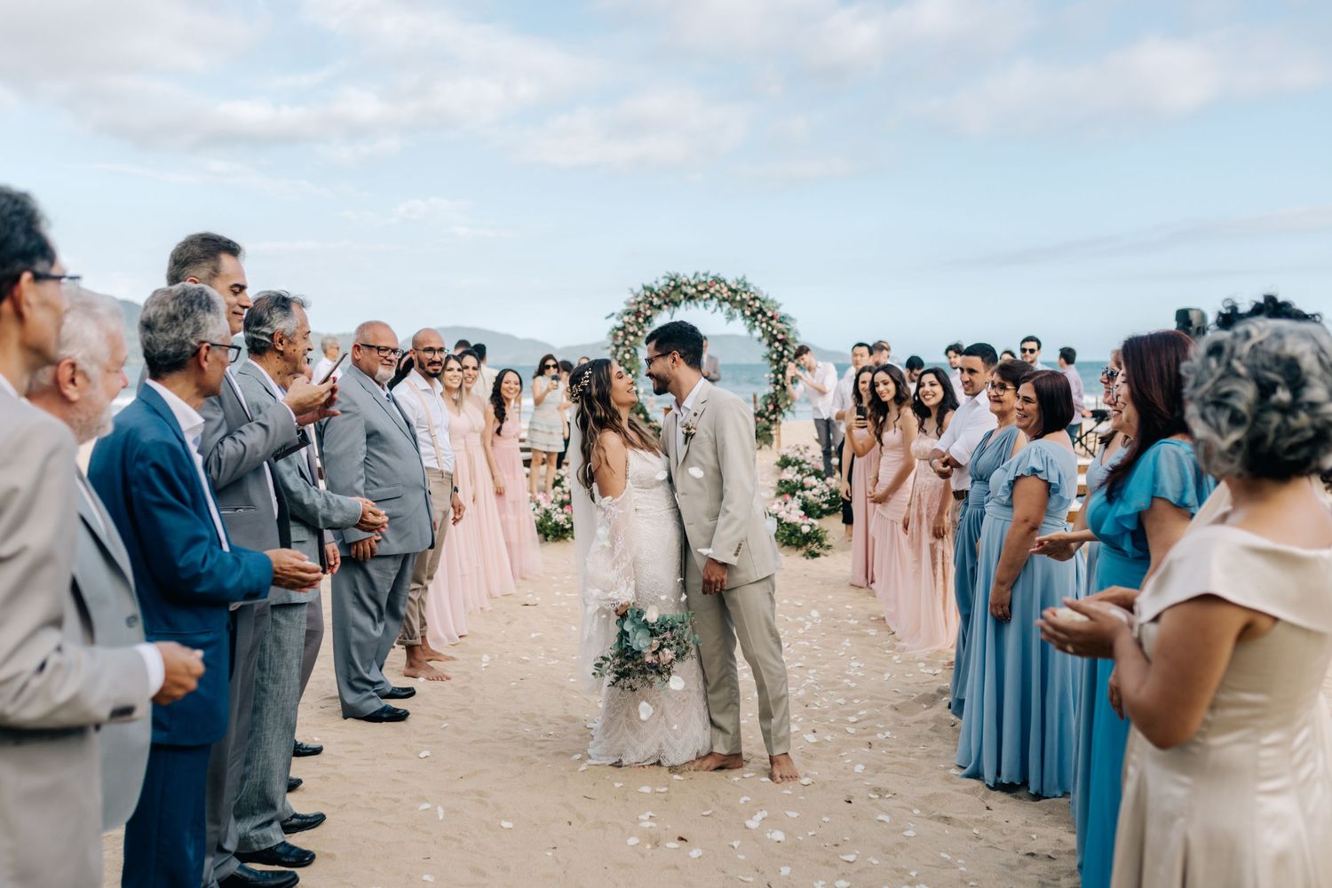 Wedding party in blue and pink attire celebrating on a scenic beach with mountains in background.
