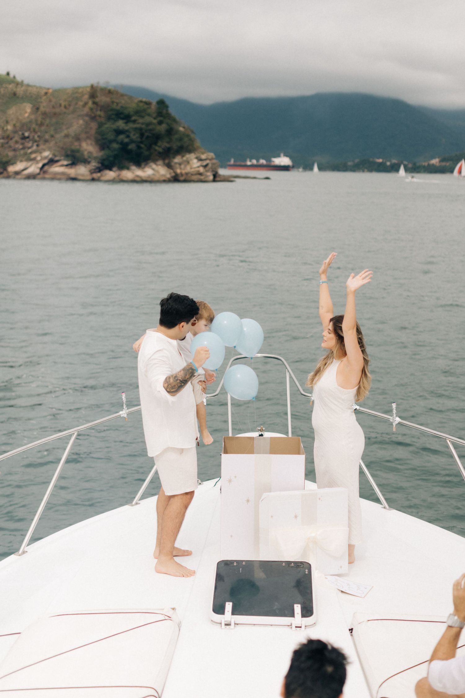 A celebration with blue balloons takes place on the bow of a yacht against a scenic harbor backdrop.