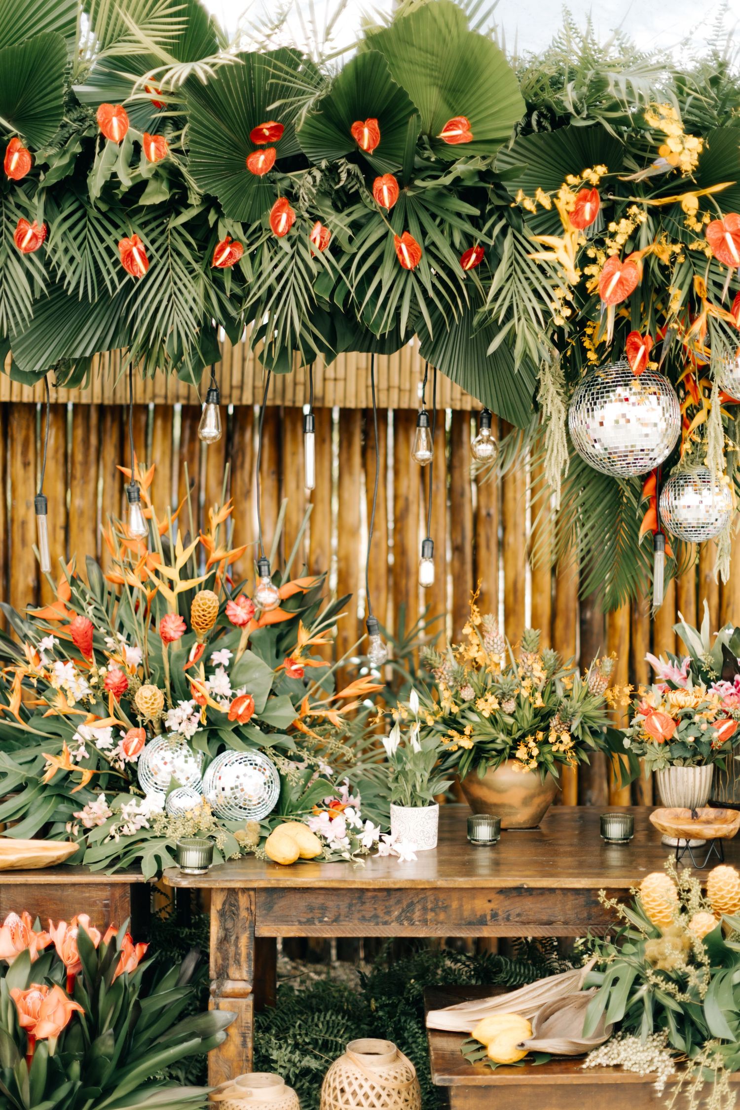Tropical wedding backdrop with palm leaves, orange flowers, and disco balls against a bamboo fence.