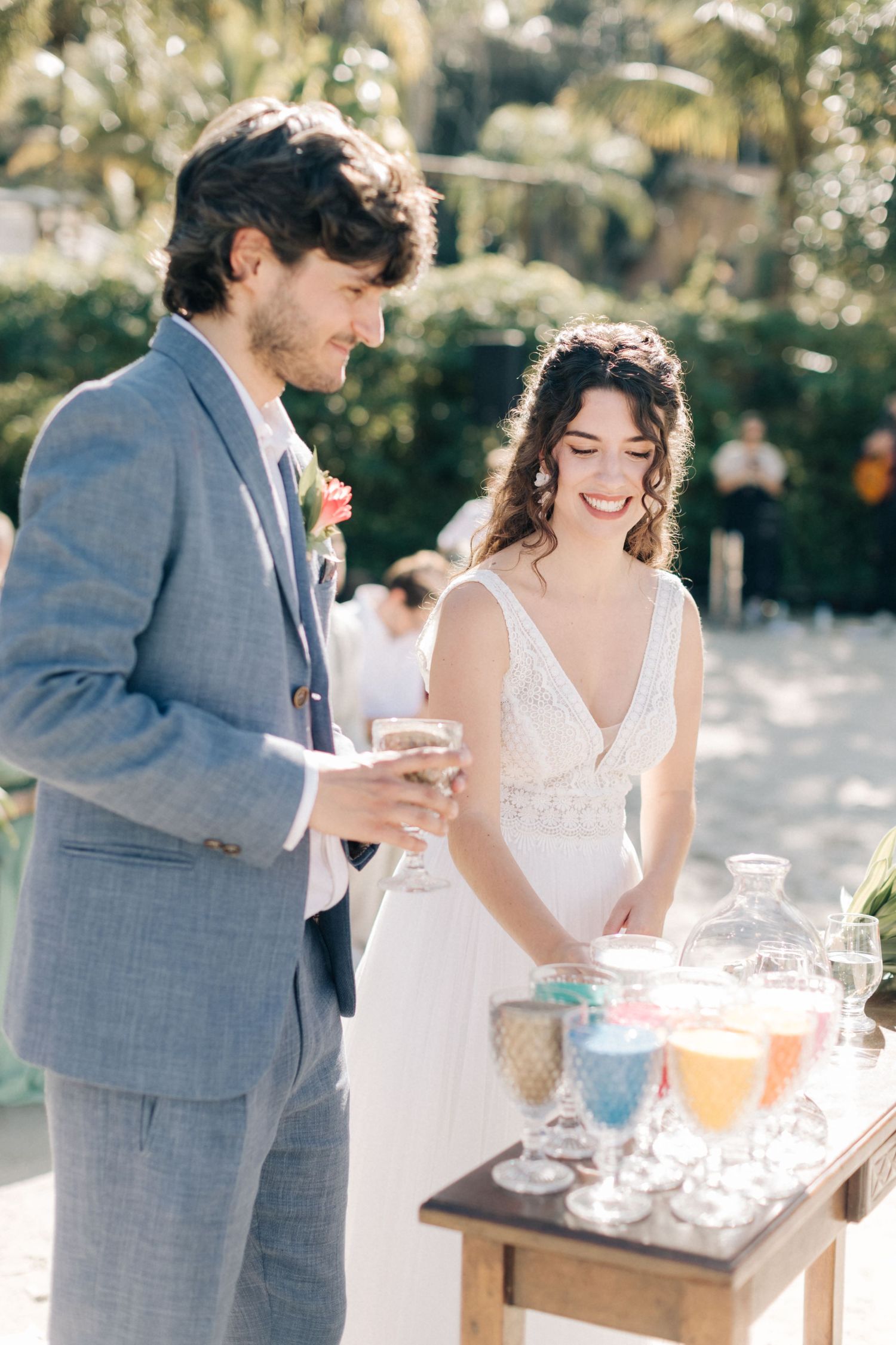Couple participates in wedding ceremony ritual next to drink station with glassware outdoors.