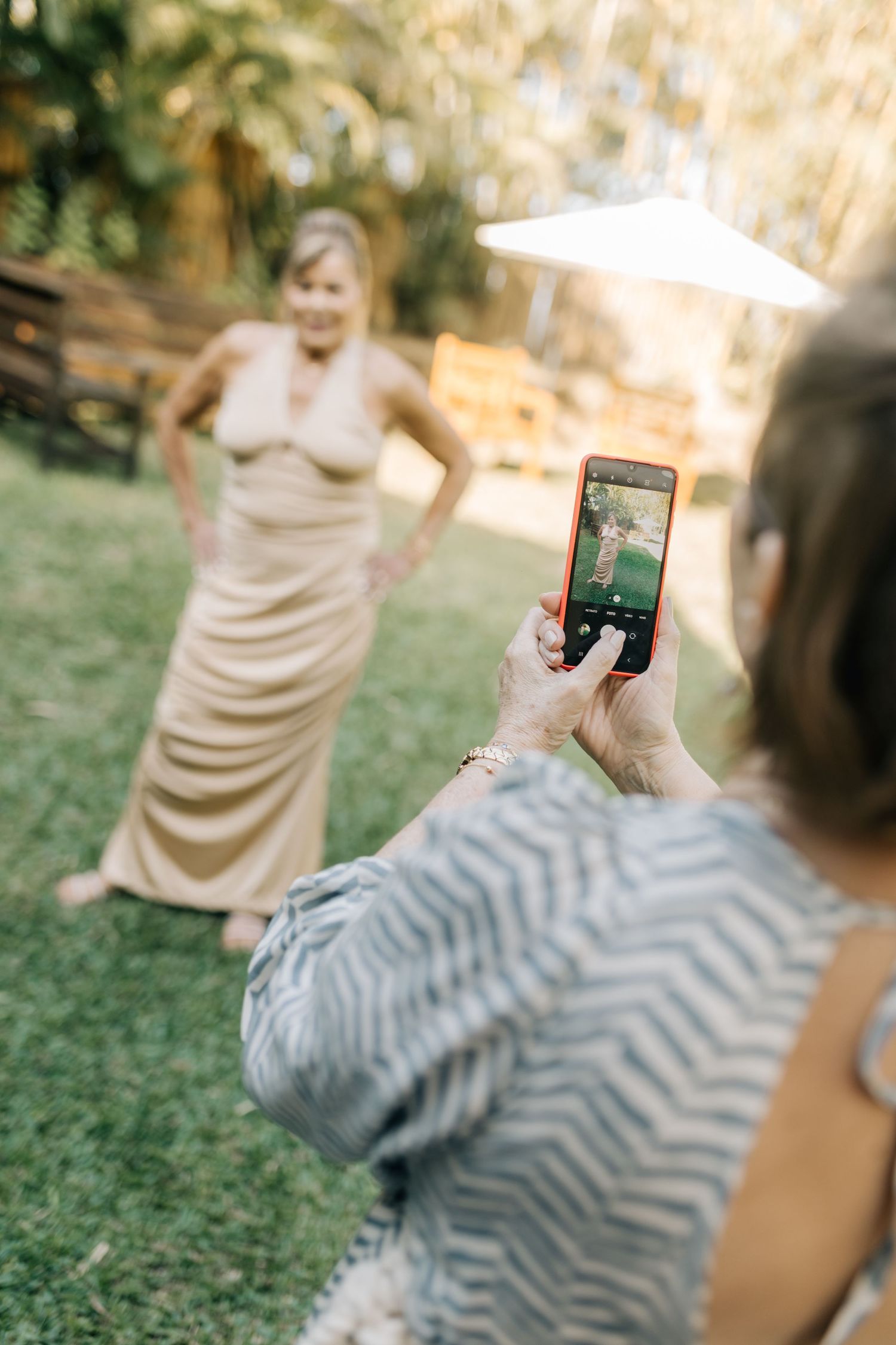 Someone takes a phone photo of a person in a champagne-colored dress on a lawn.