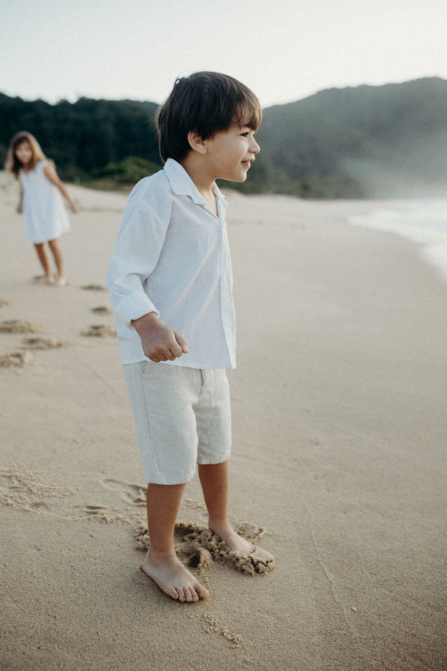 A child in white clothing standing on a misty beach with mountains in the background.