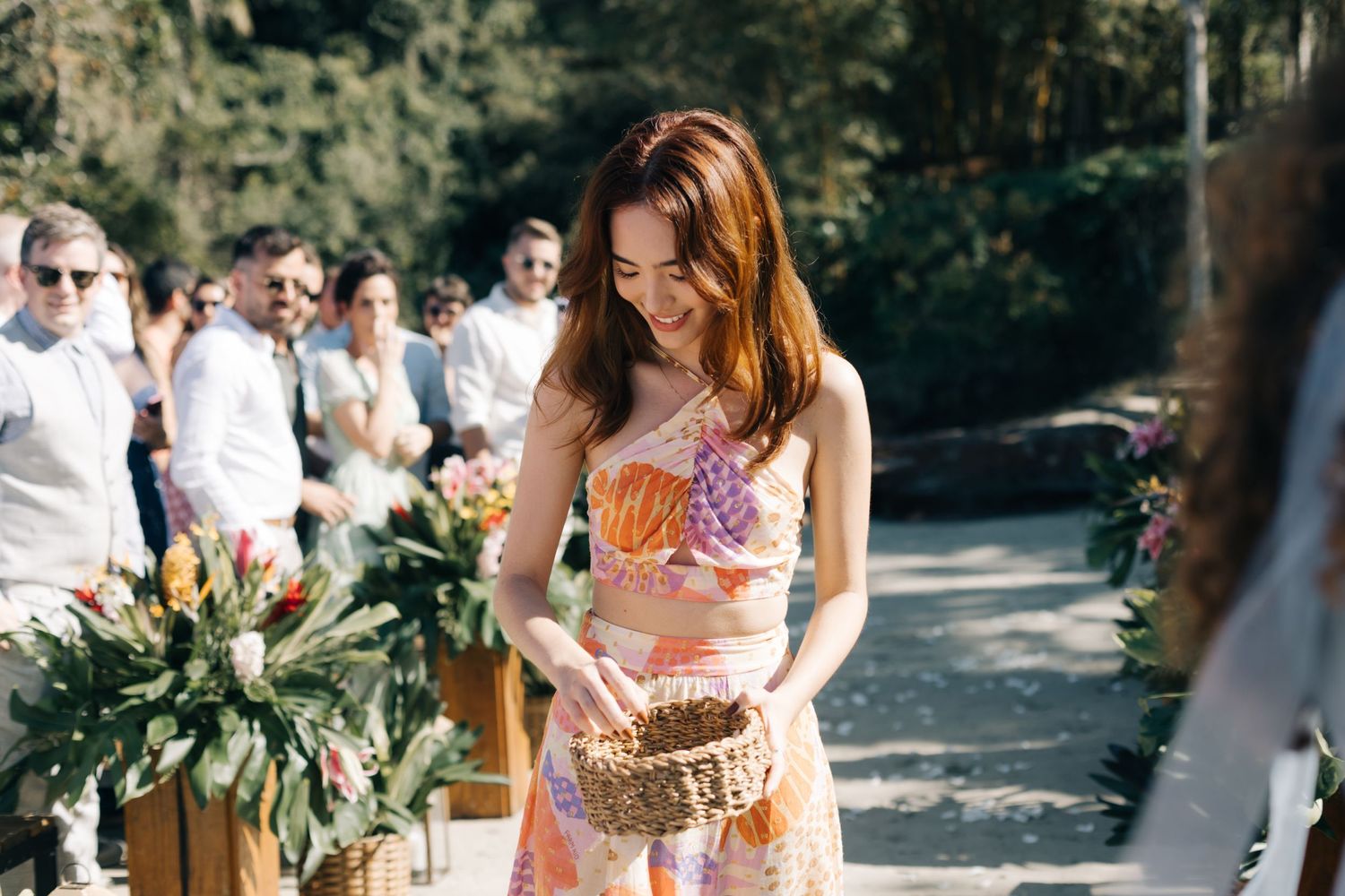 A guest in a floral dress holds a woven basket at a tropical outdoor wedding.