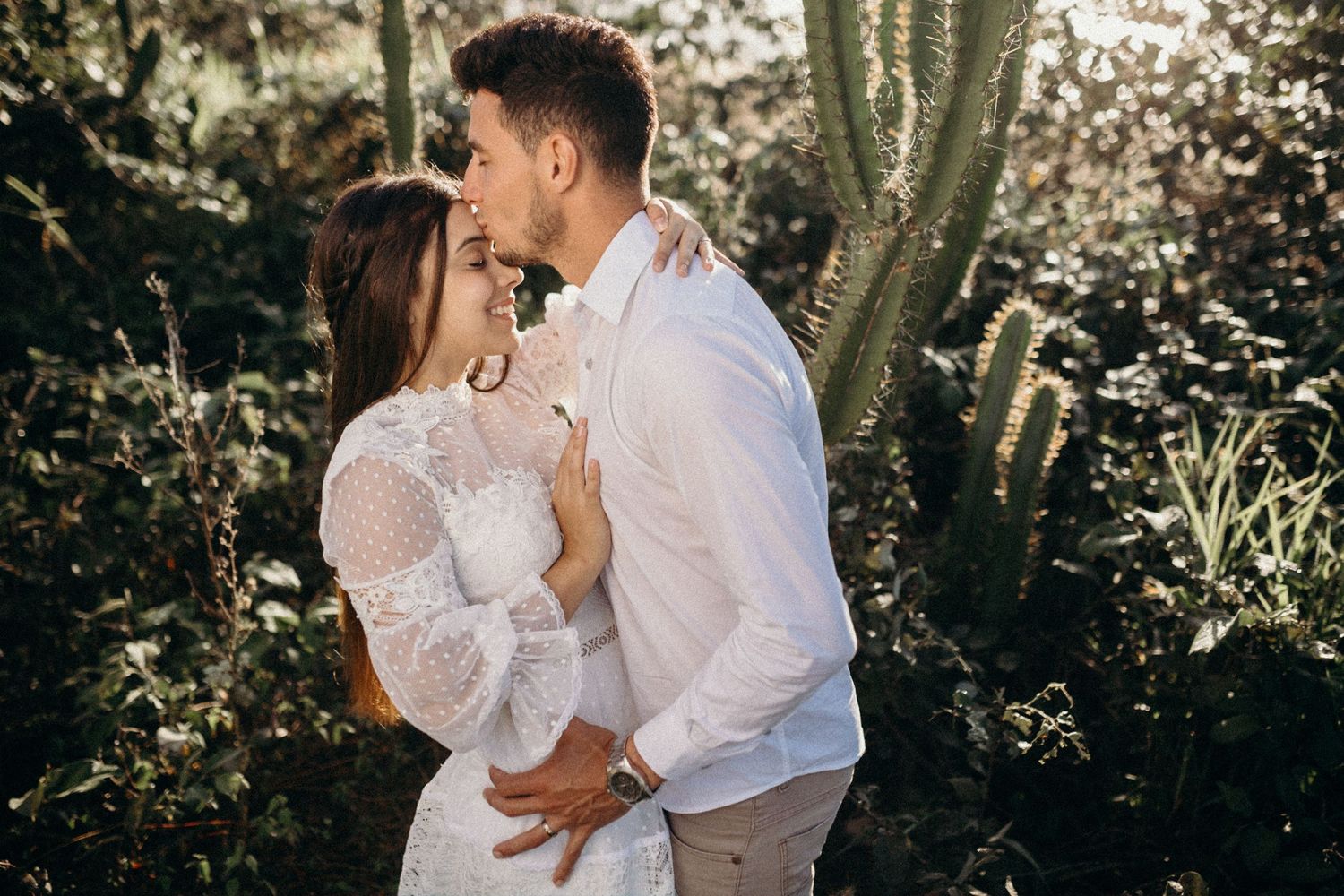 Elegant couple in white clothing share romantic embrace surrounded by desert flora.