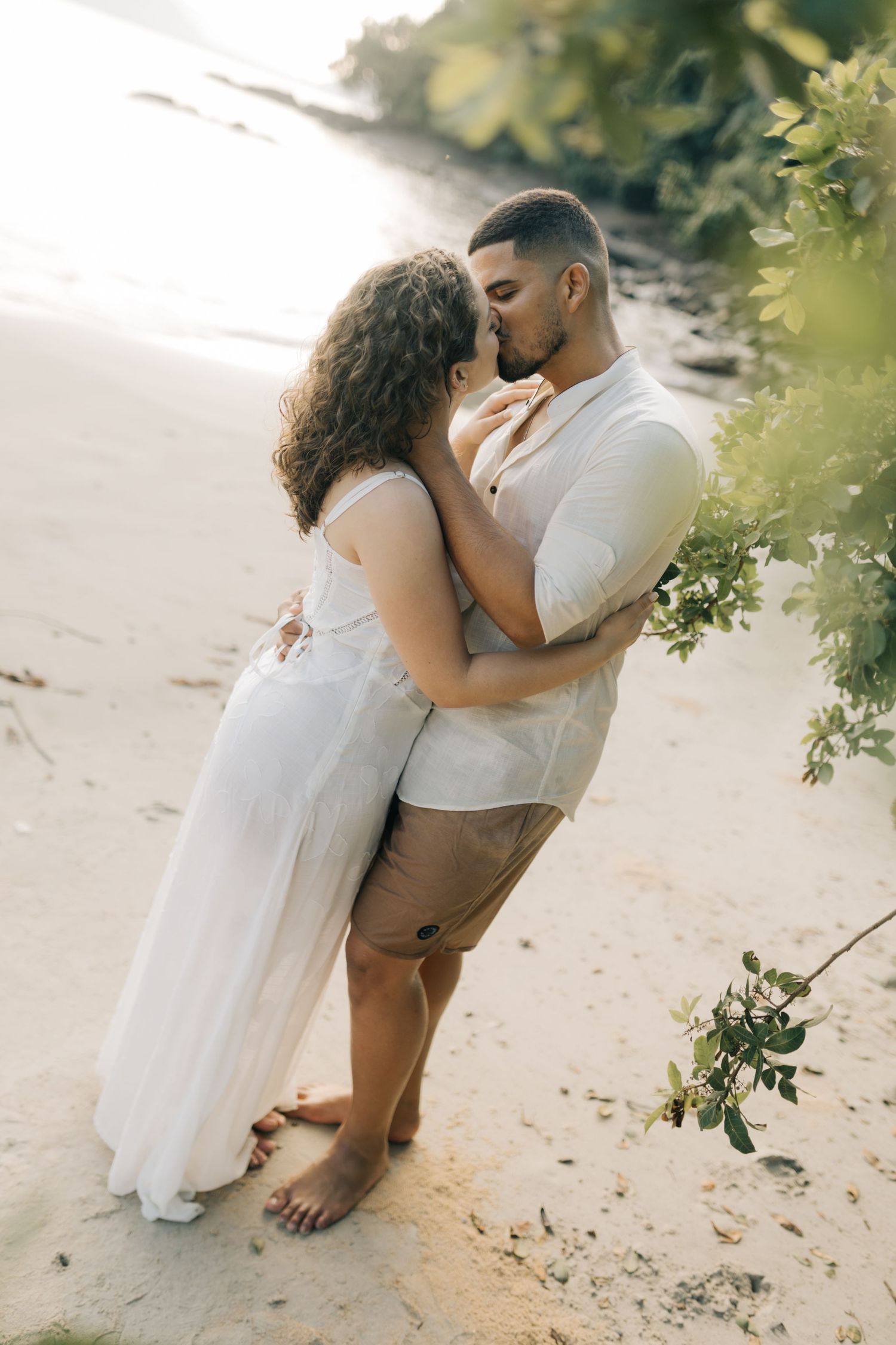 Intimate beach portrait of couple in white clothing under green leaves.