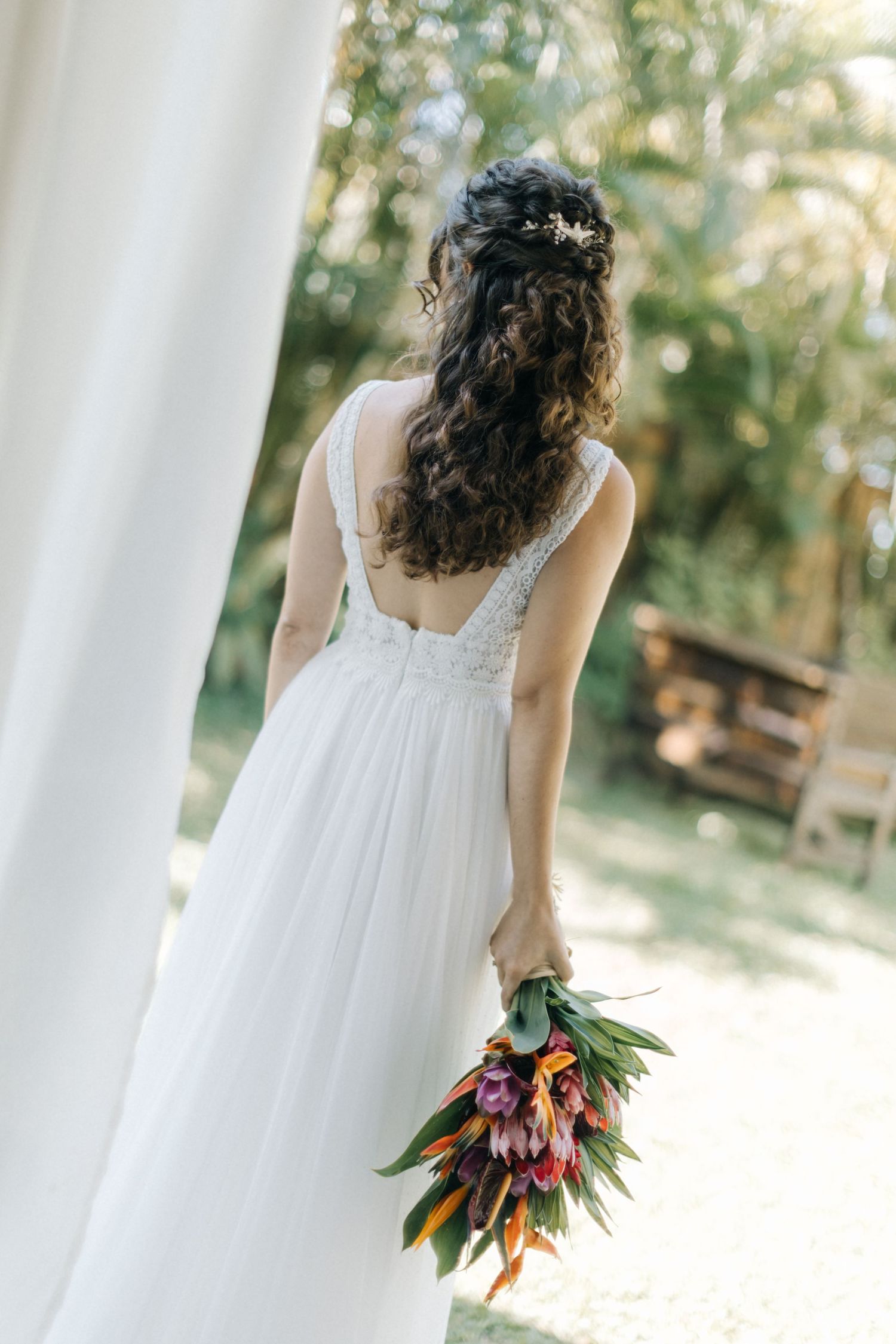 Bride in flowing white dress walks with tropical bouquet among outdoor columns.