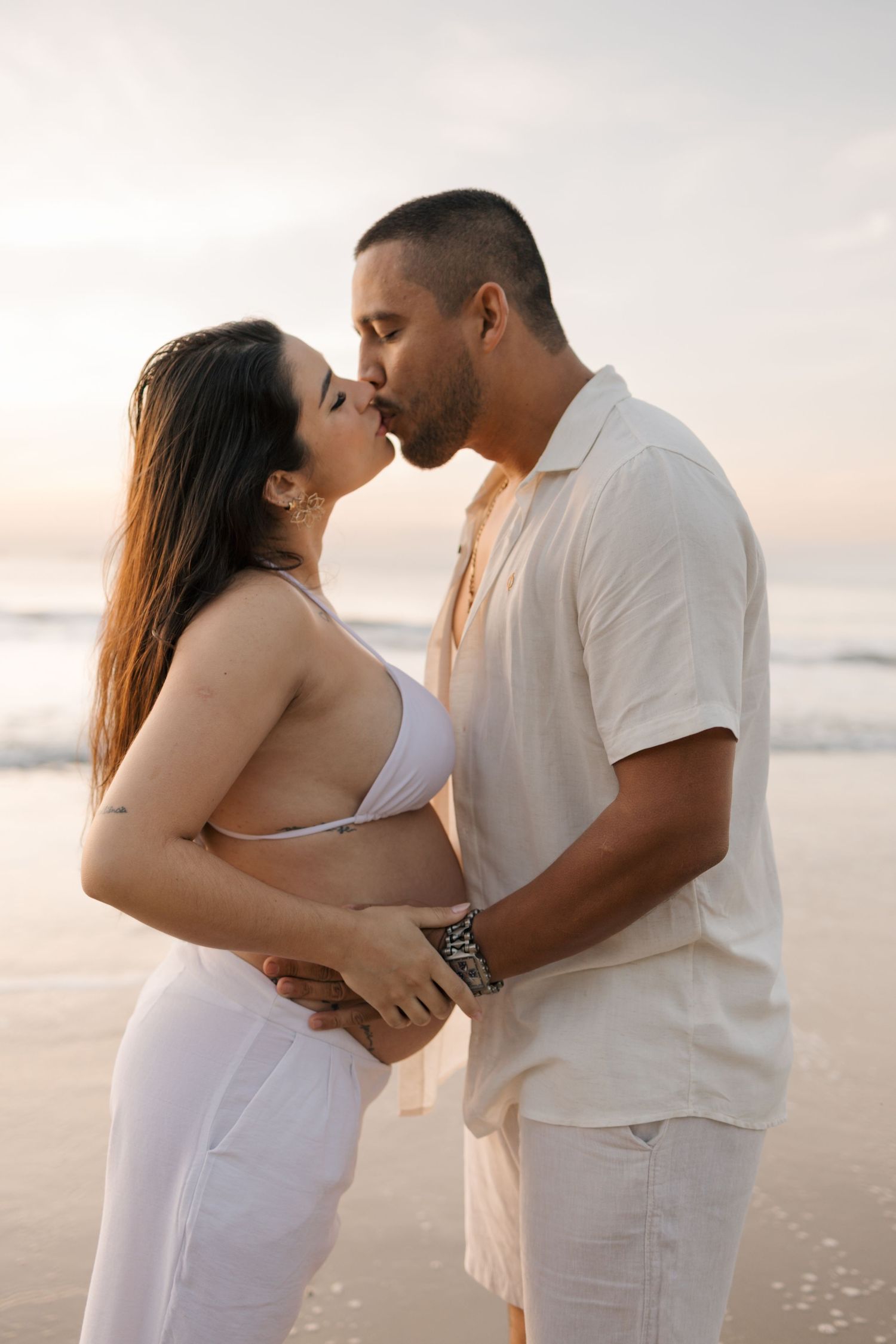 An intimate moment between a couple in white clothing standing on a beach at dusk.