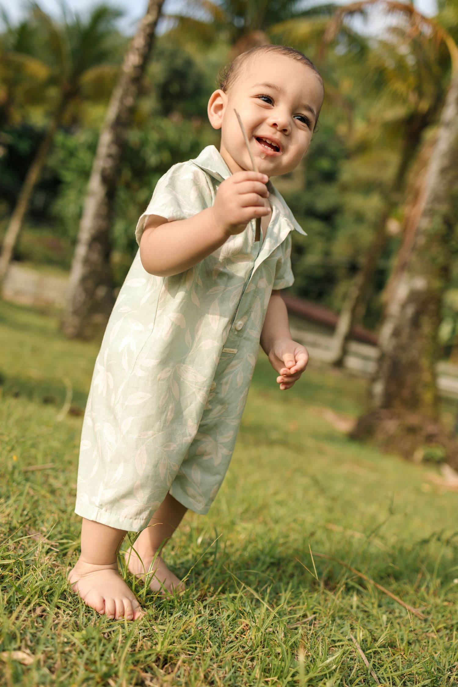 A small child wearing a light colored outfit plays and smiles while sitting on green grass with palm trees in background.