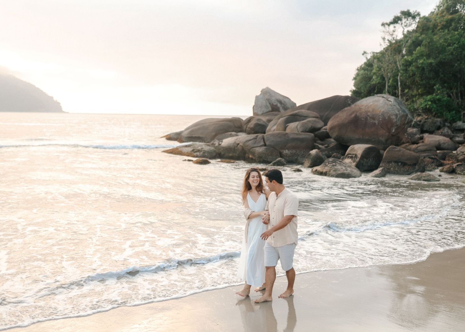 Couple walks along rocky beach shoreline during sunset photography session.