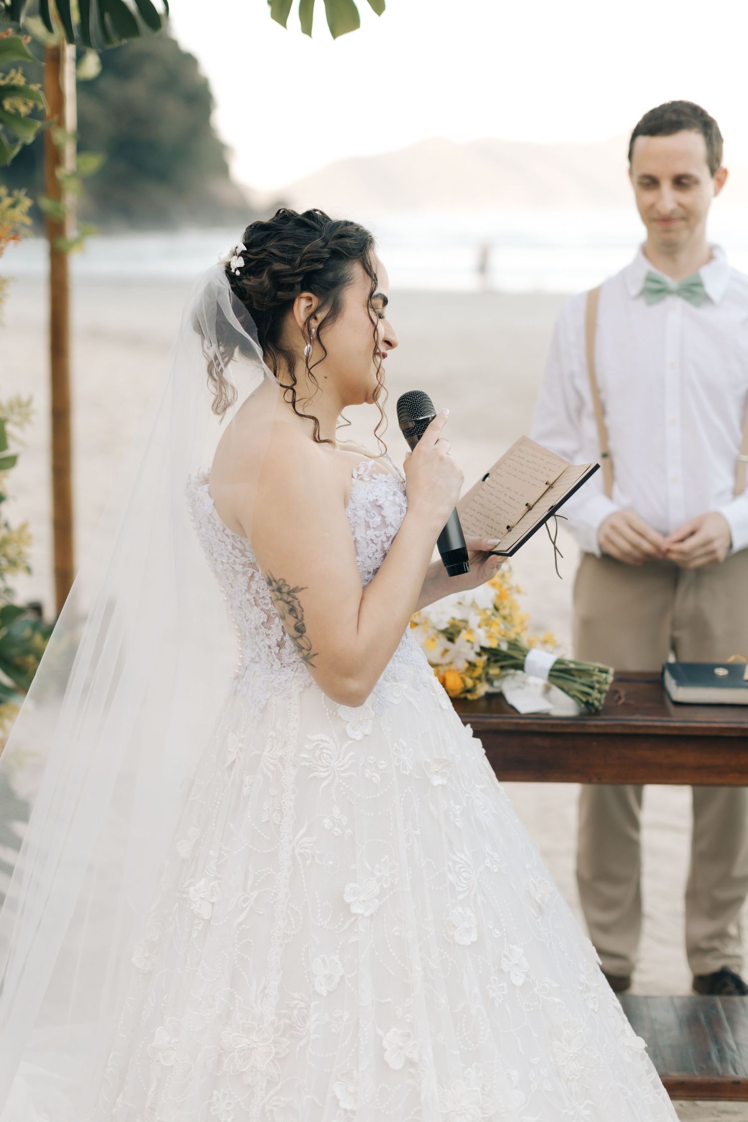 A bride in a lace white gown reads from a book during an outdoor beach wedding ceremony.