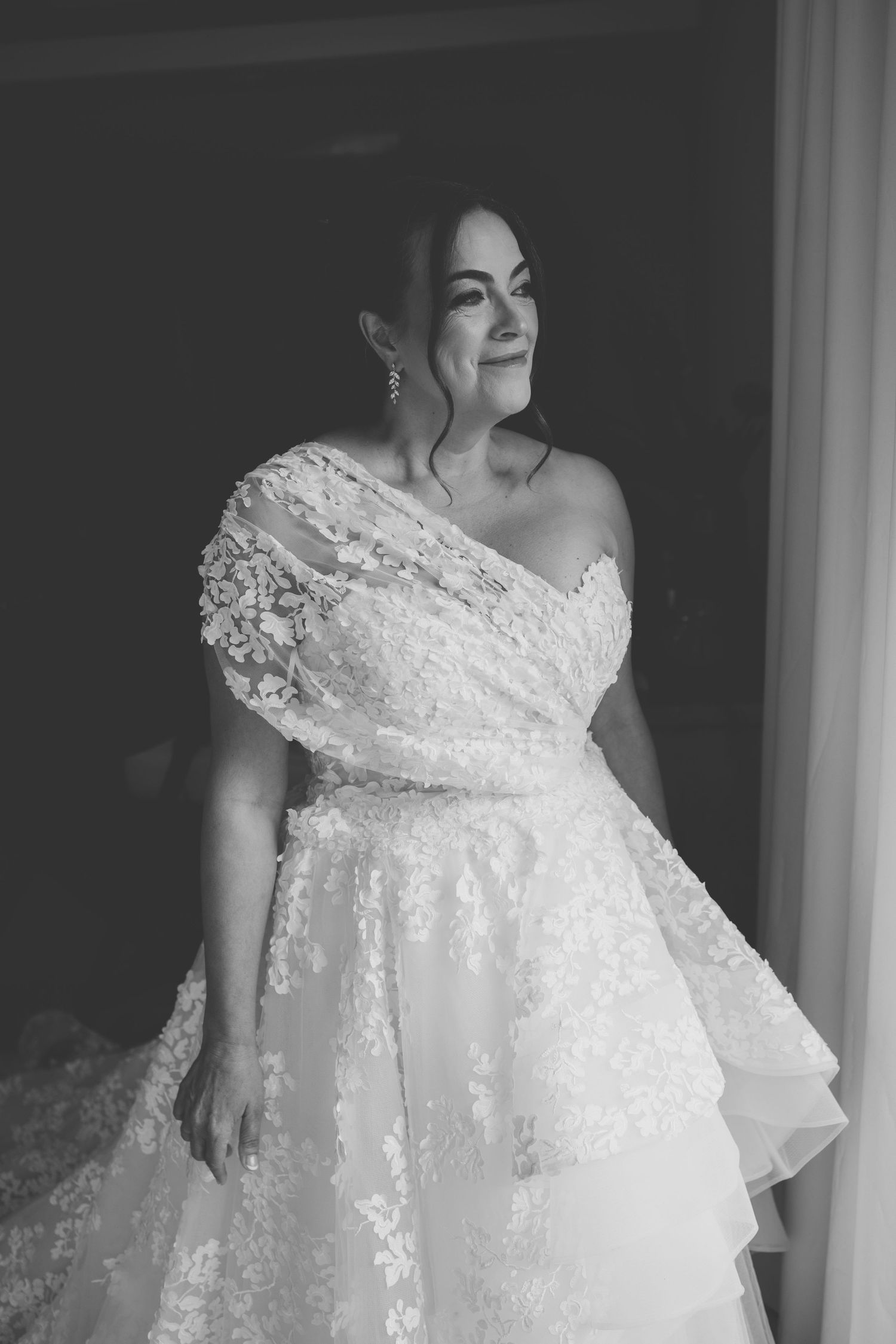 A bride in an elegant white lace wedding dress poses by a window in a black and white photograph.