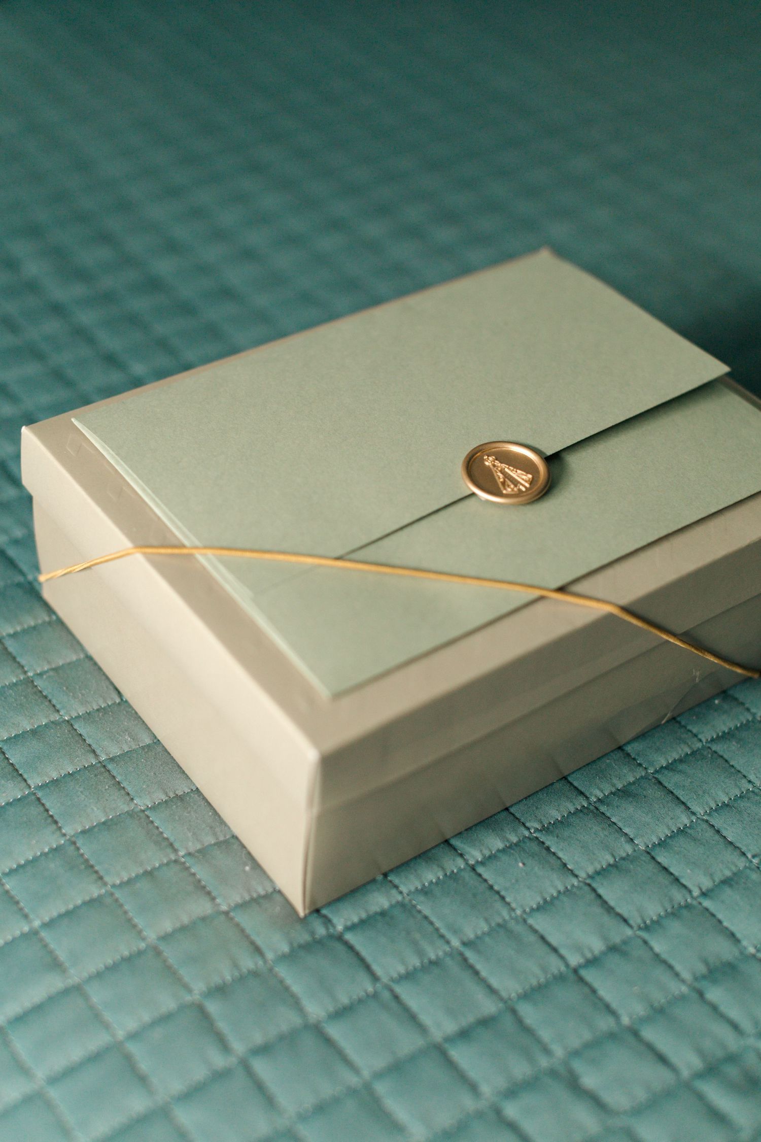 Elegant mint green gift box with gold seal and string on quilted teal surface.