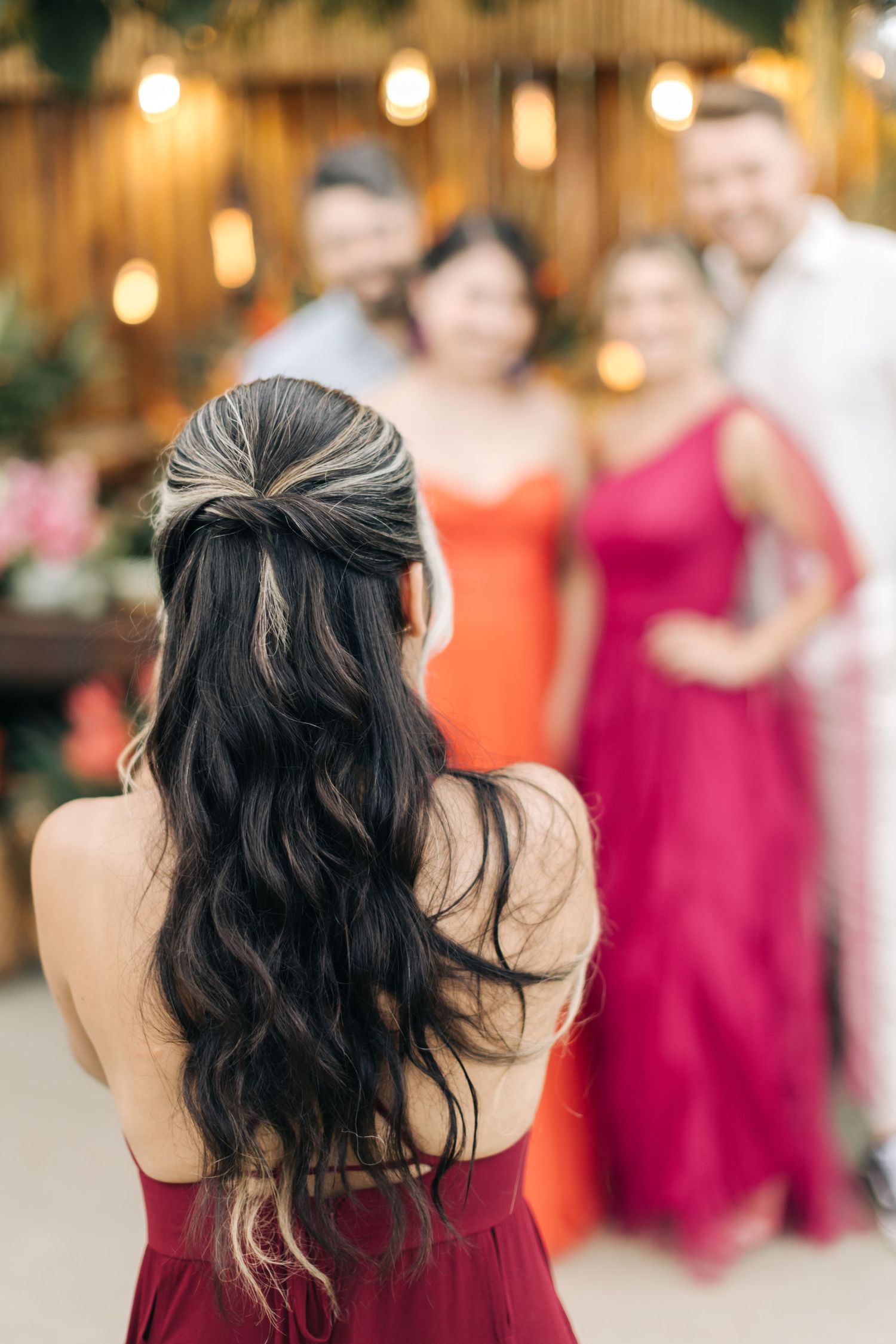 Woman in burgundy dress admires guests while taking photos at wedding venue with string lights.