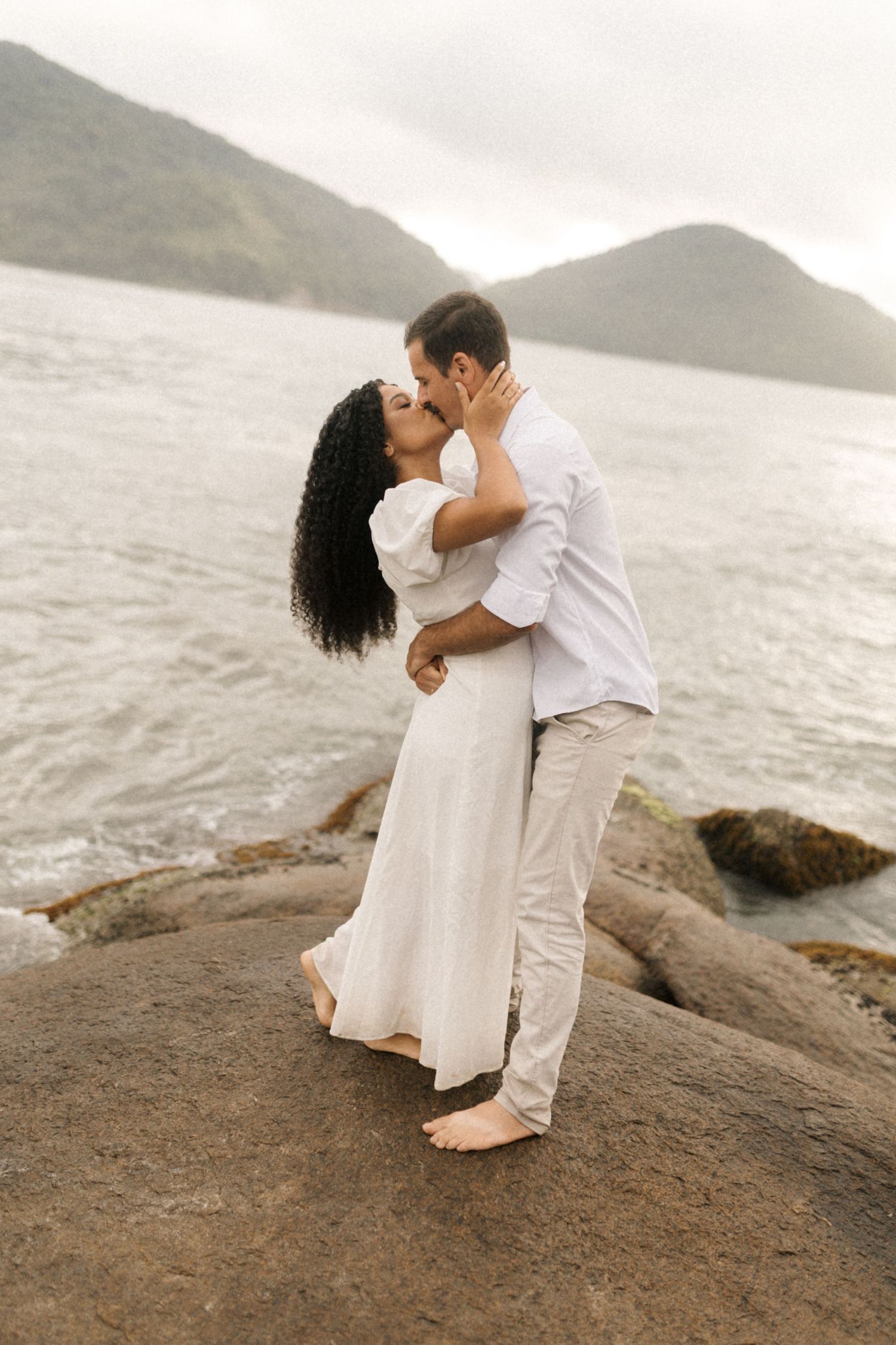 A romantic couple embraces while sitting on rocky coastal cliffs overlooking a misty ocean view.