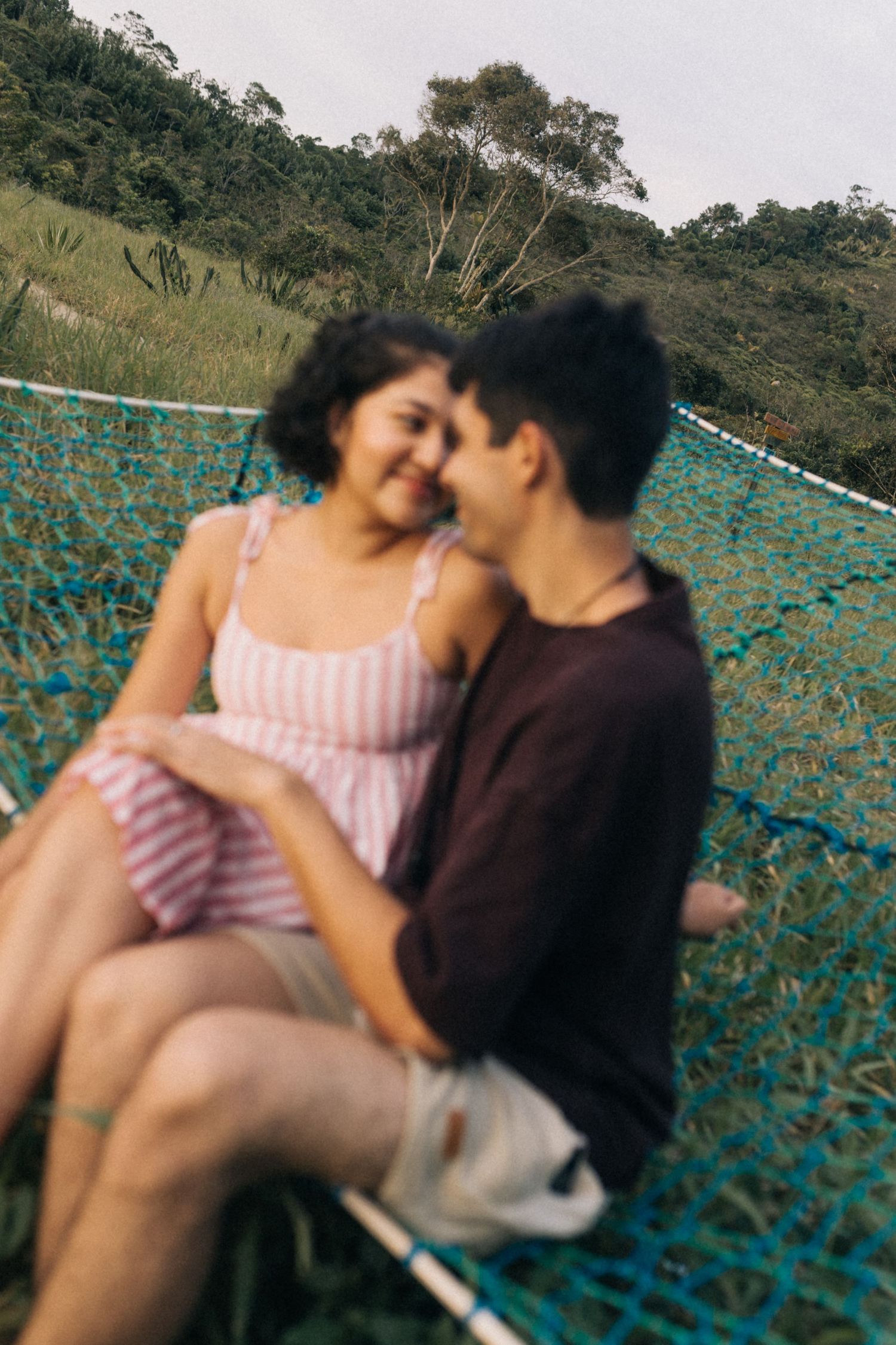 Um casal compartilha um momento íntimo enquanto está sentado em uma rede turquesa contra o cenário de uma encosta verde.