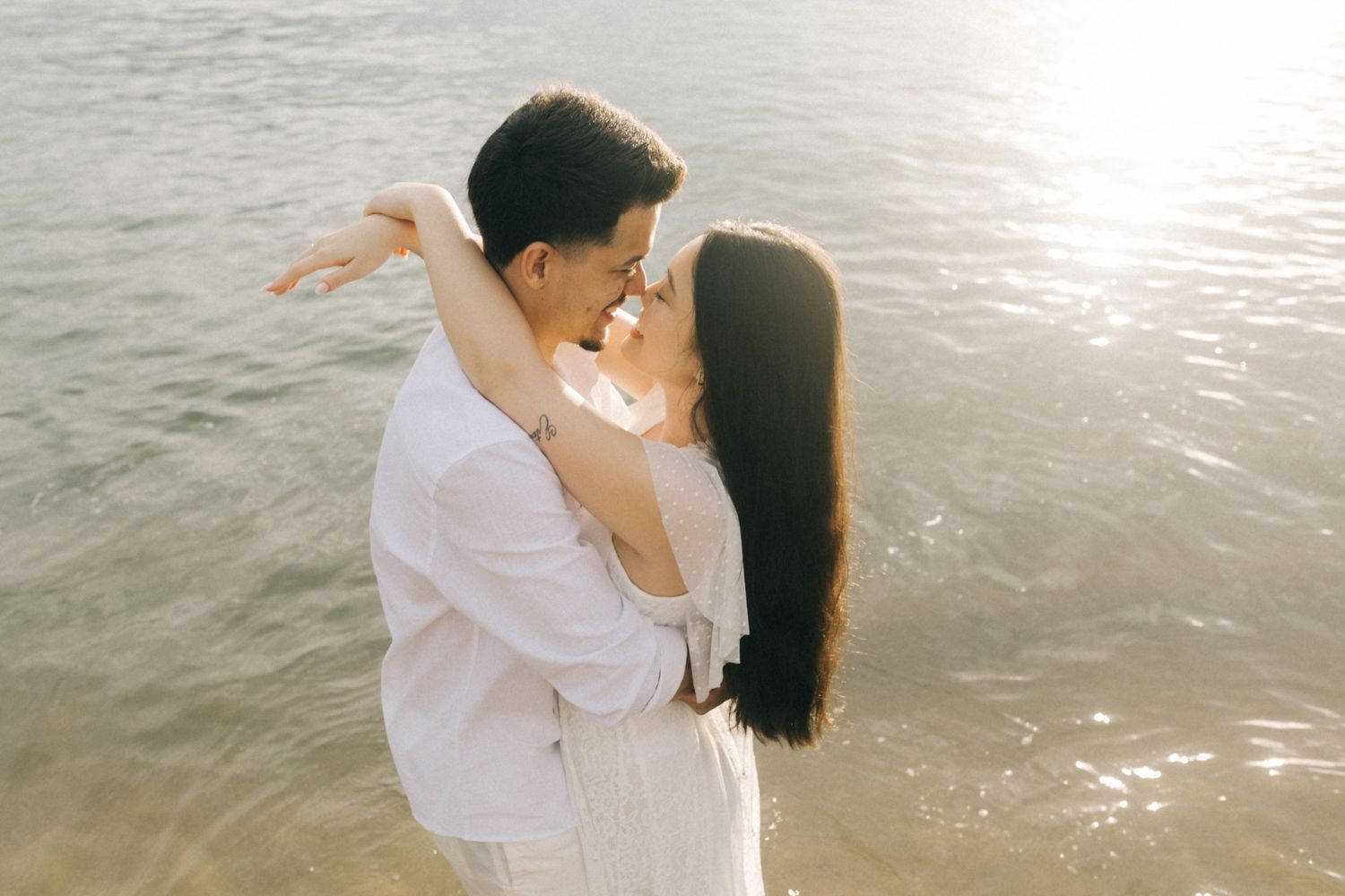 Side view of couple in white attire posing against sparkling water backdrop.