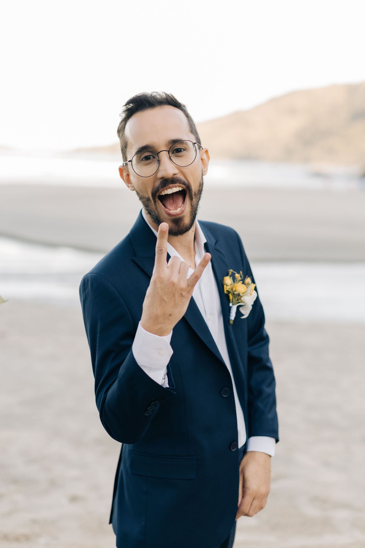 An excited person in a navy suit making a rock gesture at a beach wedding.