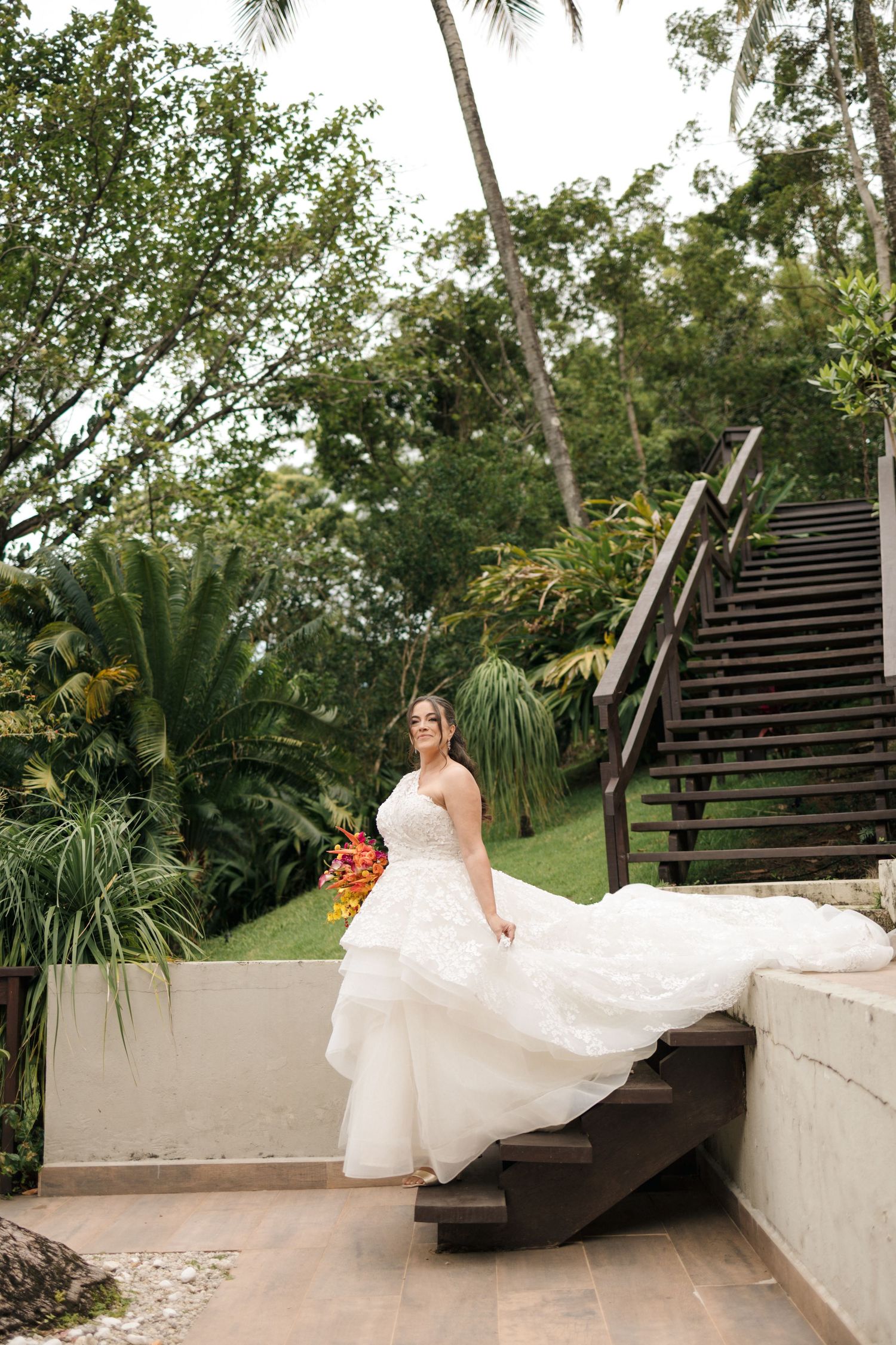 A bride in a flowing white dress poses gracefully on outdoor stone steps surrounded by tropical greenery and palm trees.