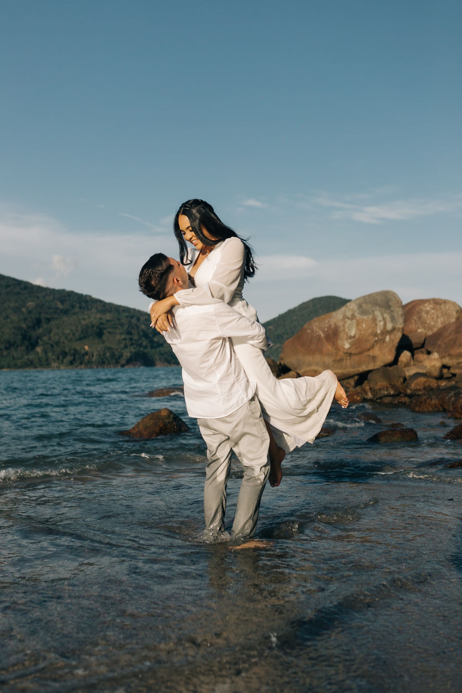 A romantic beach photo shows a playful lift pose against a mountainous backdrop.