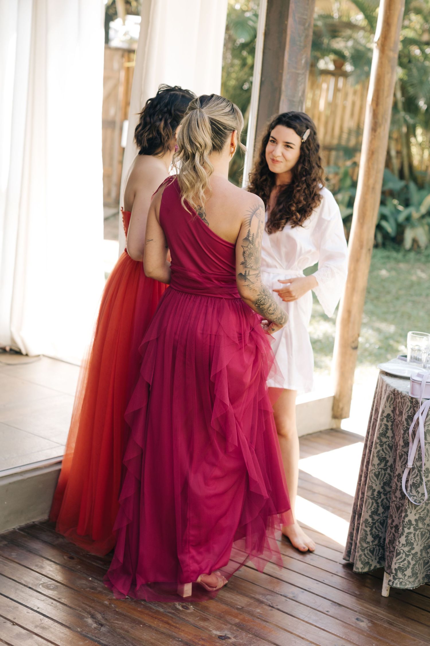 Bridesmaids in elegant red gowns chatting with person in white robe.